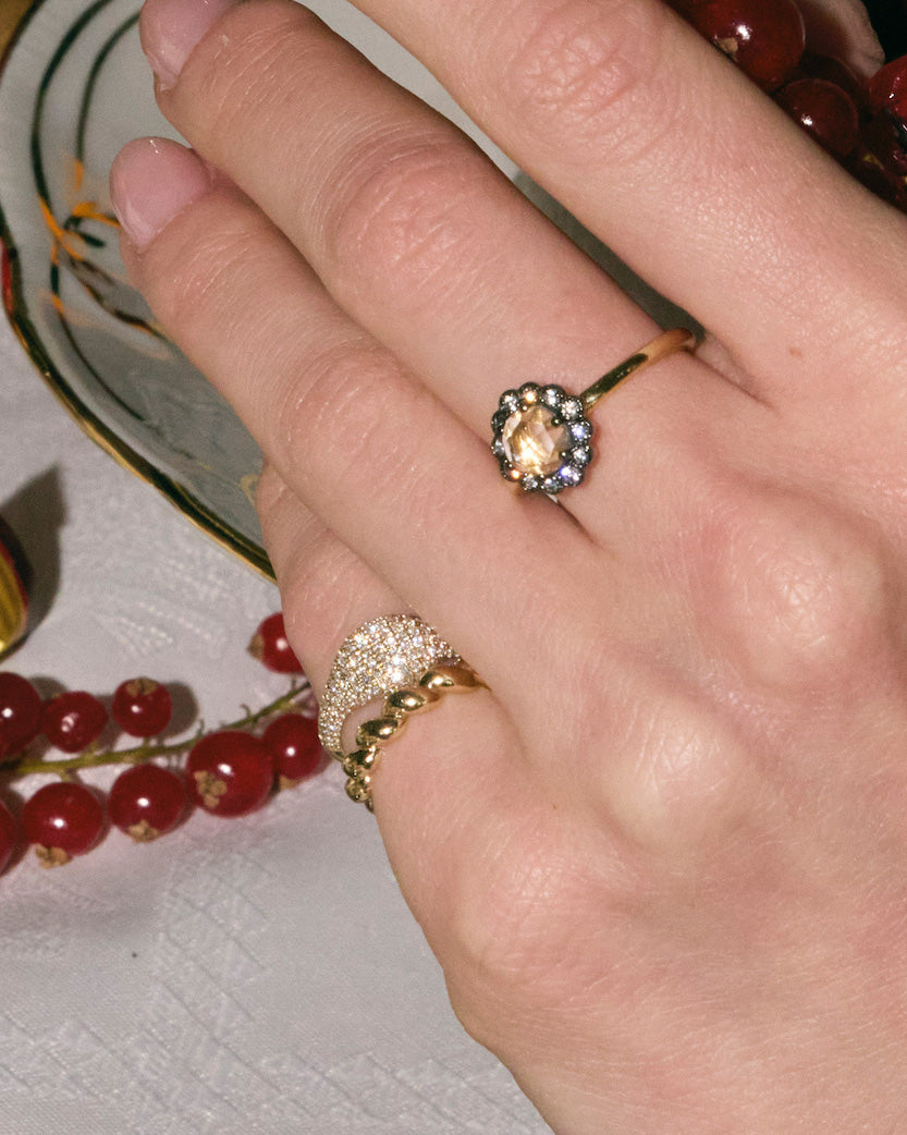 Hand wearing a gold rings with diamonds surrounded by red berries on a white surface.