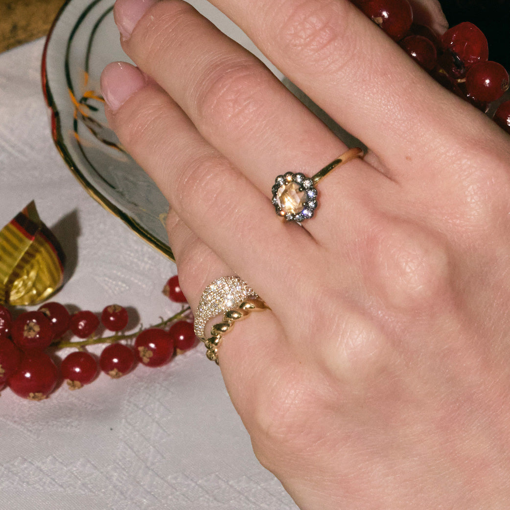 Hand wearing a gold rings with diamonds surrounded by red berries on a white surface.