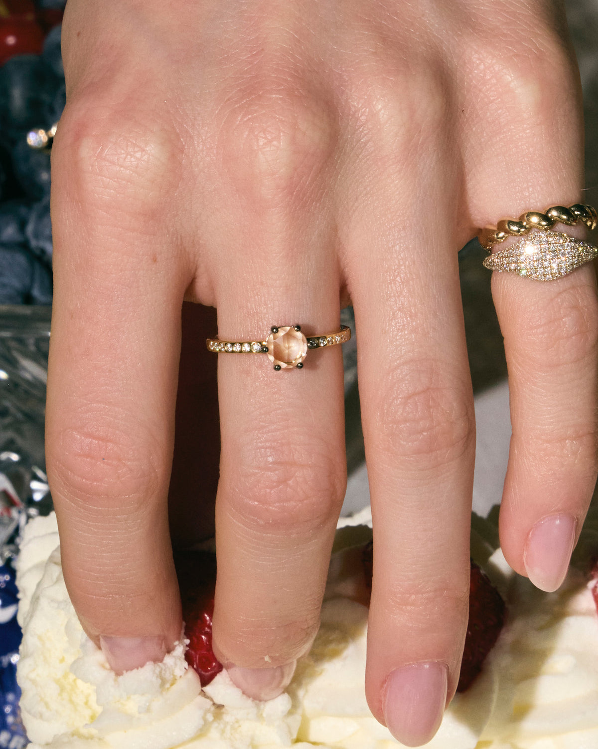 Close-up of a hand wearing three rings, one with rose-cut diamond, a pinky paved diamonds ring and a twisted band, on a dessert background