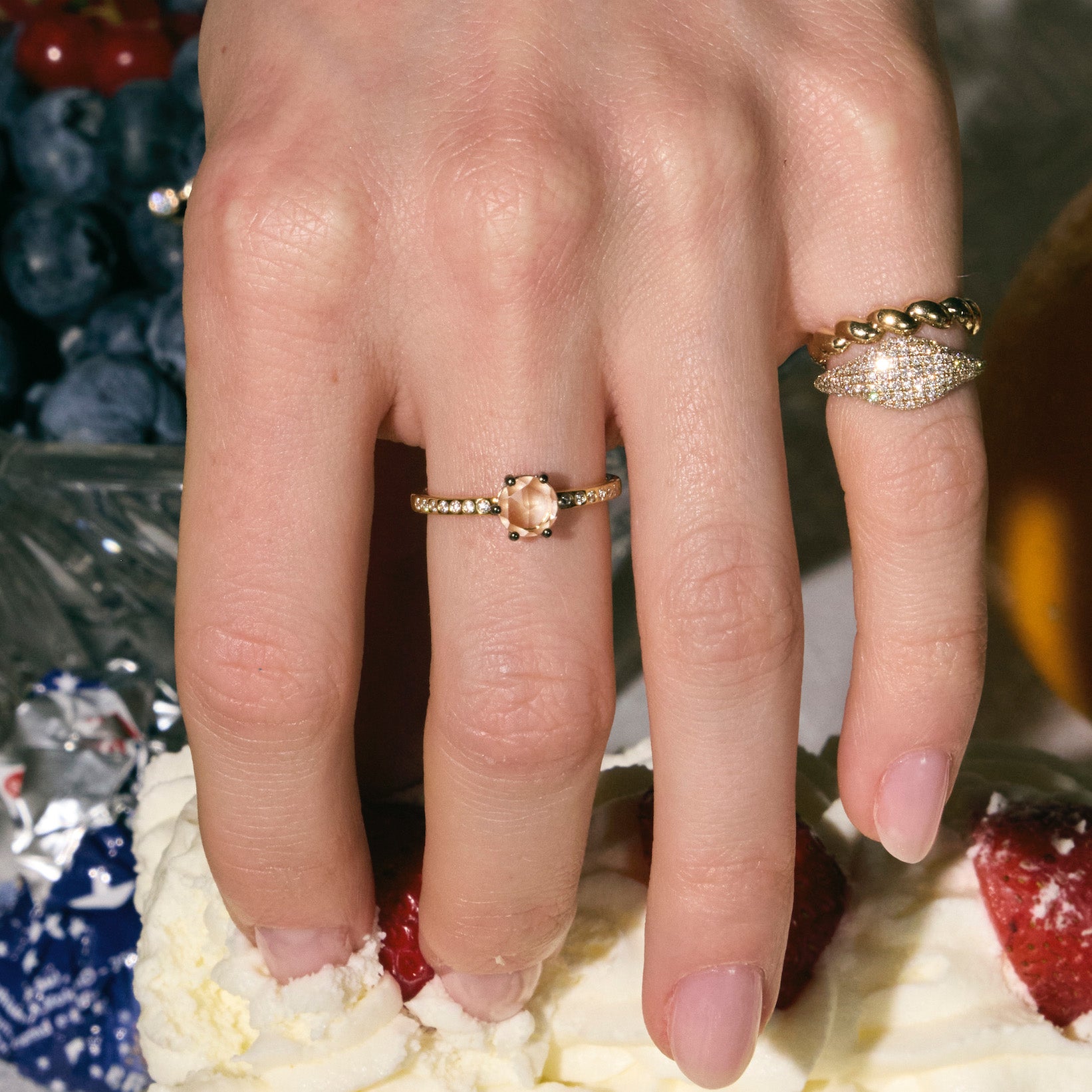 Close-up of a hand wearing three rings, one with rose-cut diamond, a pinky paved diamonds ring and a twisted band, on a dessert background