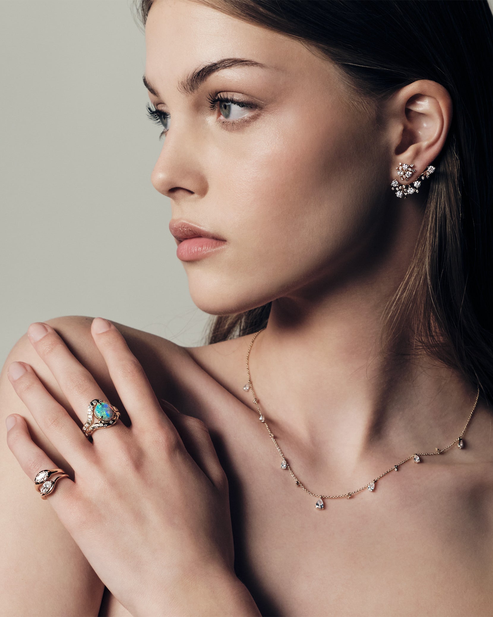 A woman wearing a ring with a australian opal and diamonds, two pinky ring with marquise diamonds set in curvy rose gold bands. She uis also wearing a teardrop diaomonds necklace and a cluster diamonds earcrawler earring.
