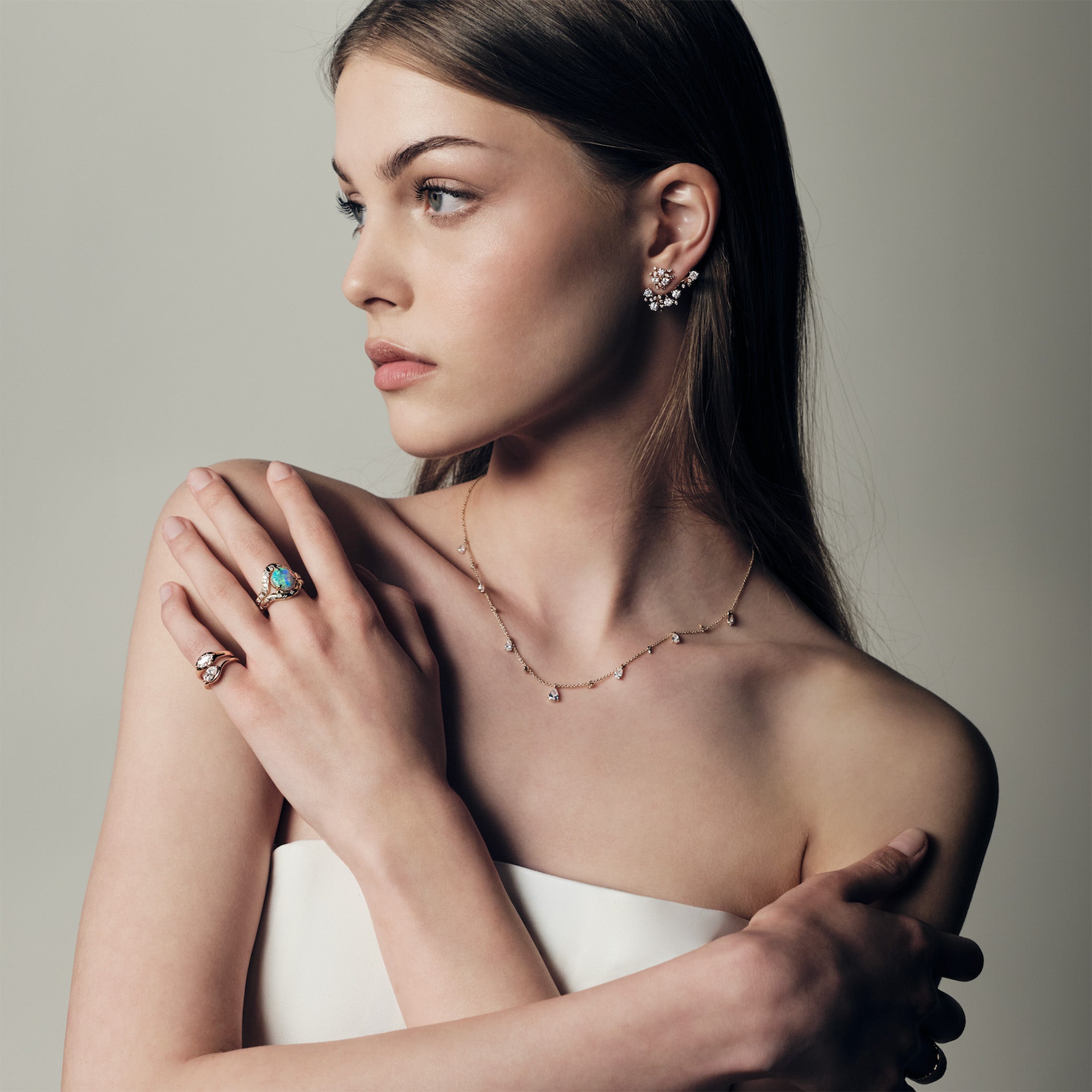 A woman wearing a ring with a australian opal and diamonds, two pinky ring with marquise diamonds set in curvy rose gold bands. She uis also wearing a teardrop diaomonds necklace and a cluster diamonds earcrawler earring.
