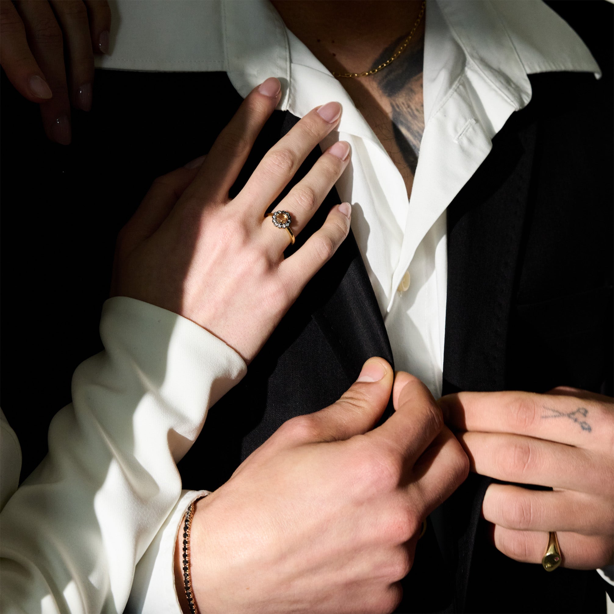 a couple having an intimate moment dressed in their wedding attire. He is wearing a black suit, a gold chain, a pinky signet ring and a black diamonds tennis bracelet, and she is wearing a white dress and a gorgeous vintage style ring