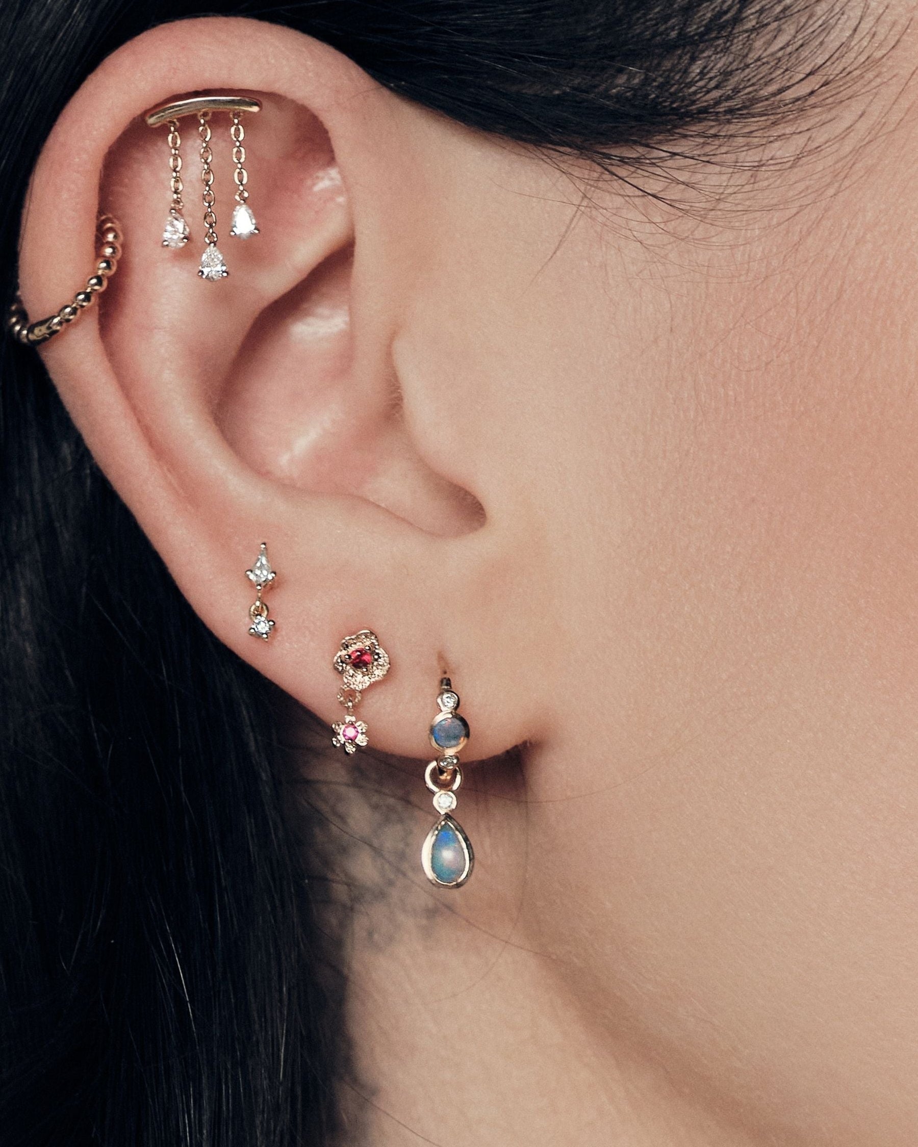 Close-up of an ear with multiple earrings on a blurred background. Chandelier diamonds stud, bubble hoop, Floating Kite Diamond stud, Wild Rose Stud With Ruby, Tiny Dew Drops Hoops and Teardrop Australian Opals Earrings accessories.