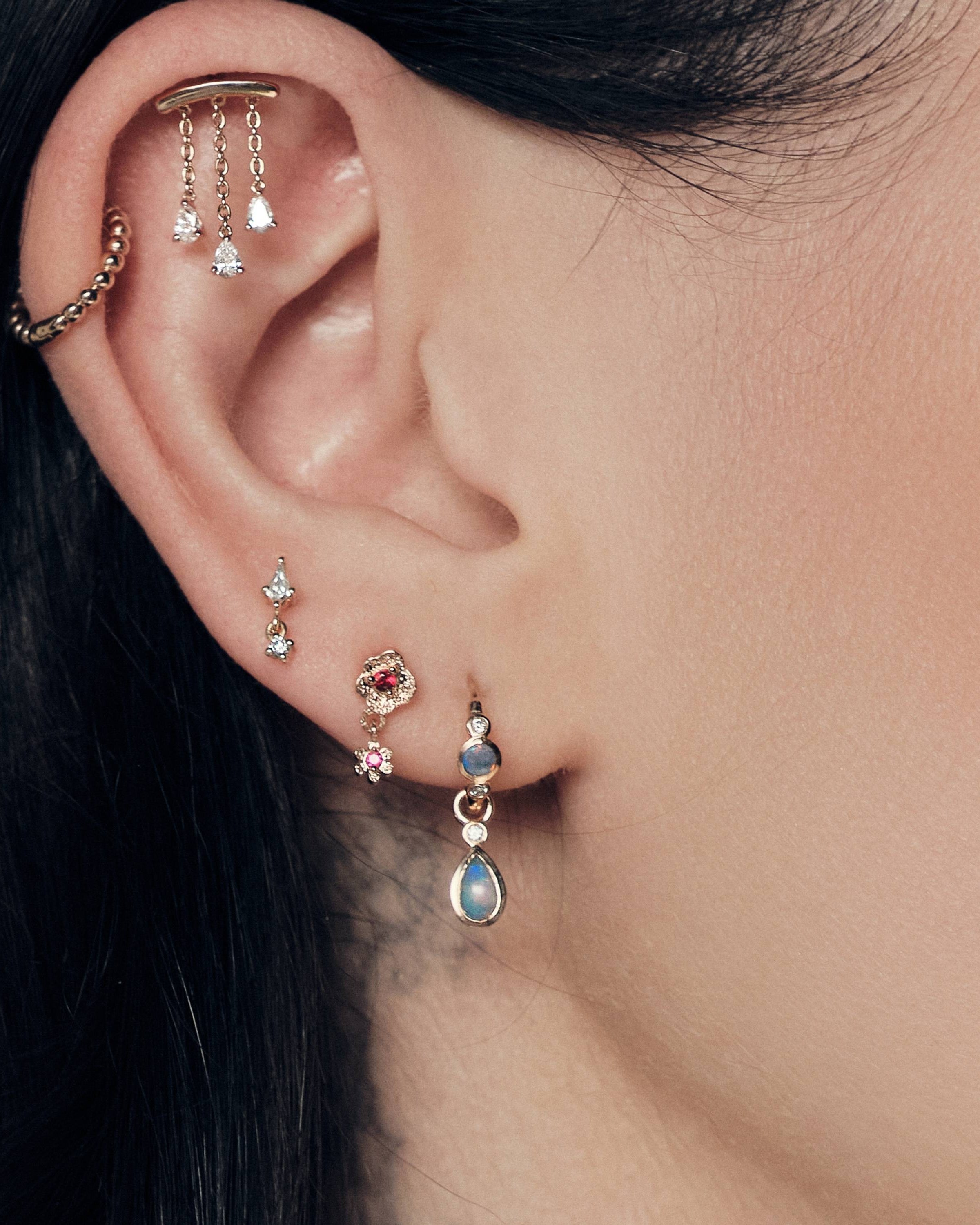 Close-up of an ear with The Chandelier Stud, Bubble Time 13mm Hoops, Floating Kite Diamond Stud, The Wild Rose Ruby Stud, Tiny Dew Drops Australian Opal Hoops, focusing on jewelry details.
