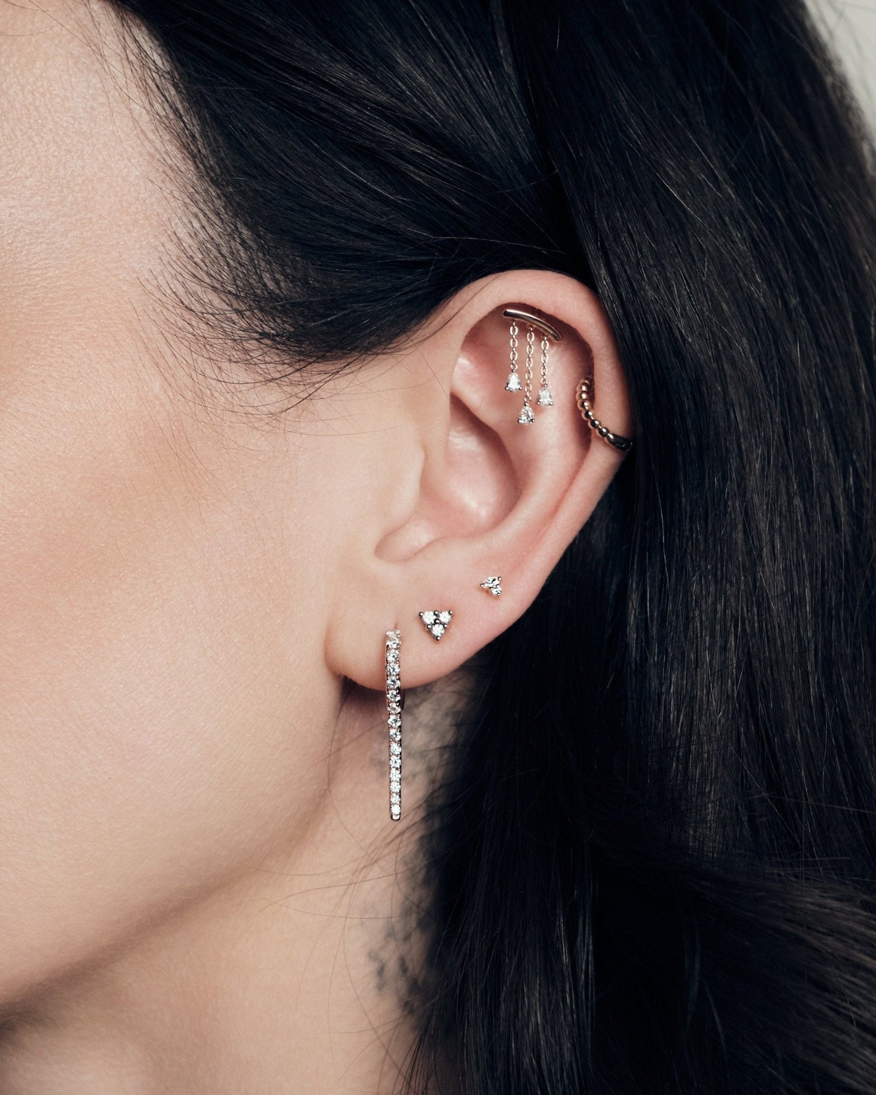 Close-up of an ear with multiple earrings, chandelier teardrop diamonds stud, bubble time 13mm hoop, marquise and round diamond stud, dot dot dot, icicle earring, against a neutral background