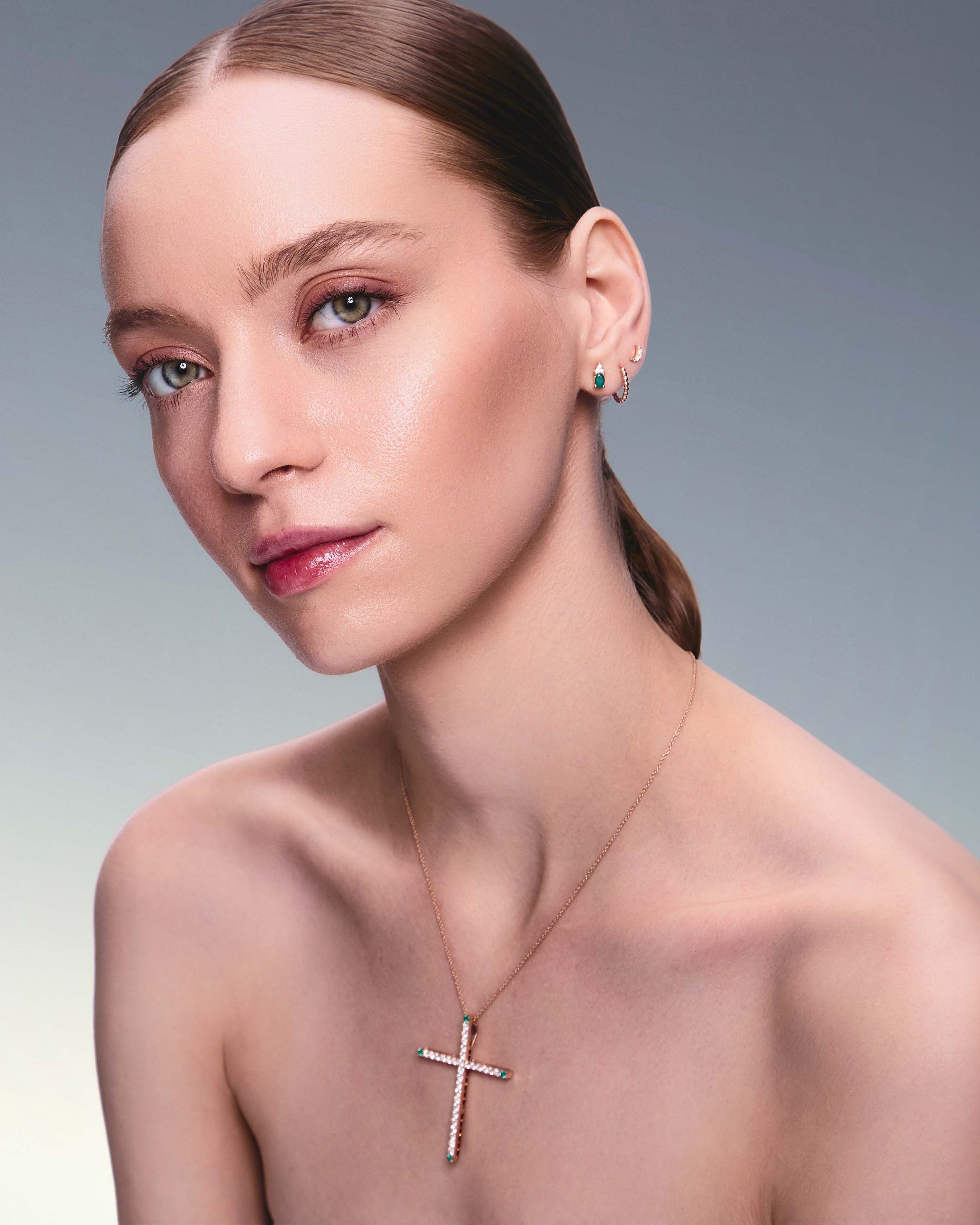 A person wearing a gold cross pendant necklace with white diamonds and emeralds.