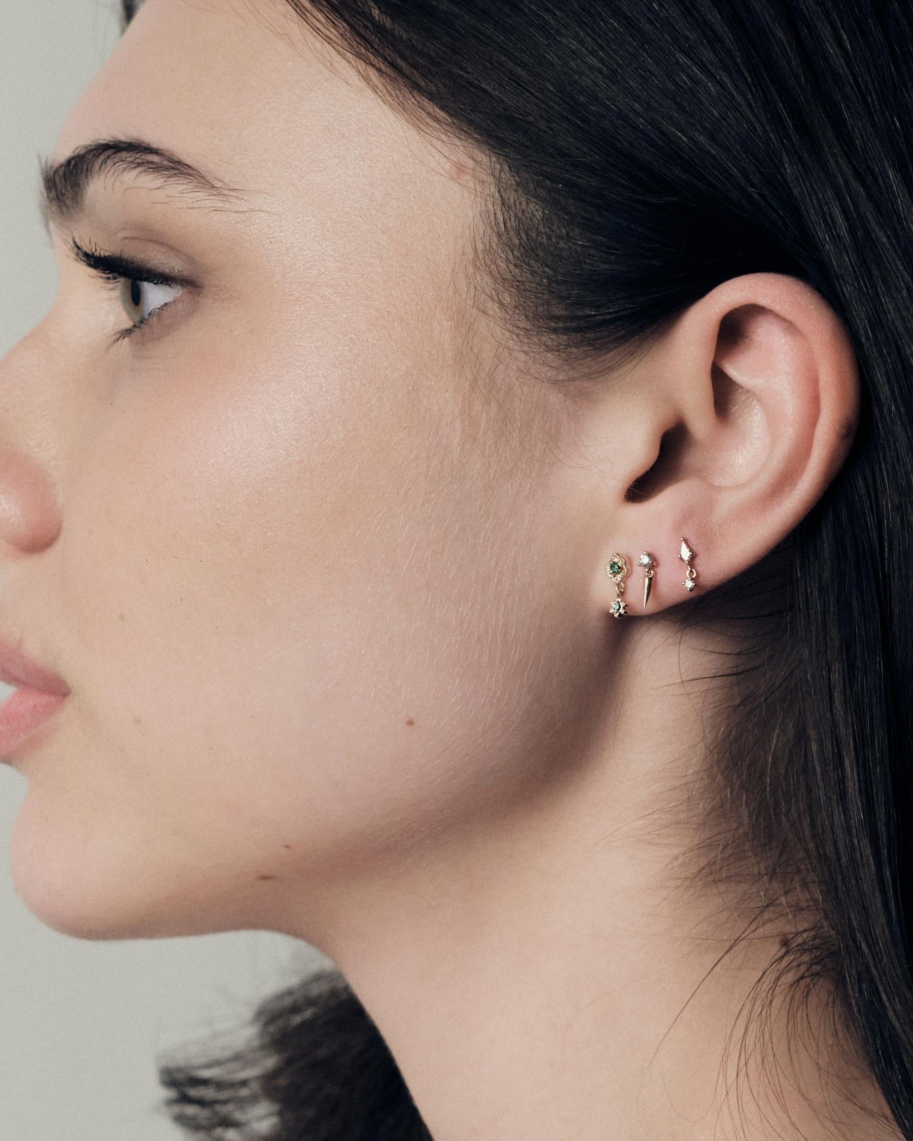 Close-up of a woman's profile wearing earrings on a neutral background. She is wearing Wild Rose Gold studs with emeralds, Floating Gold icicle Stud and Floating Kite Stud madeof yellow gold.
