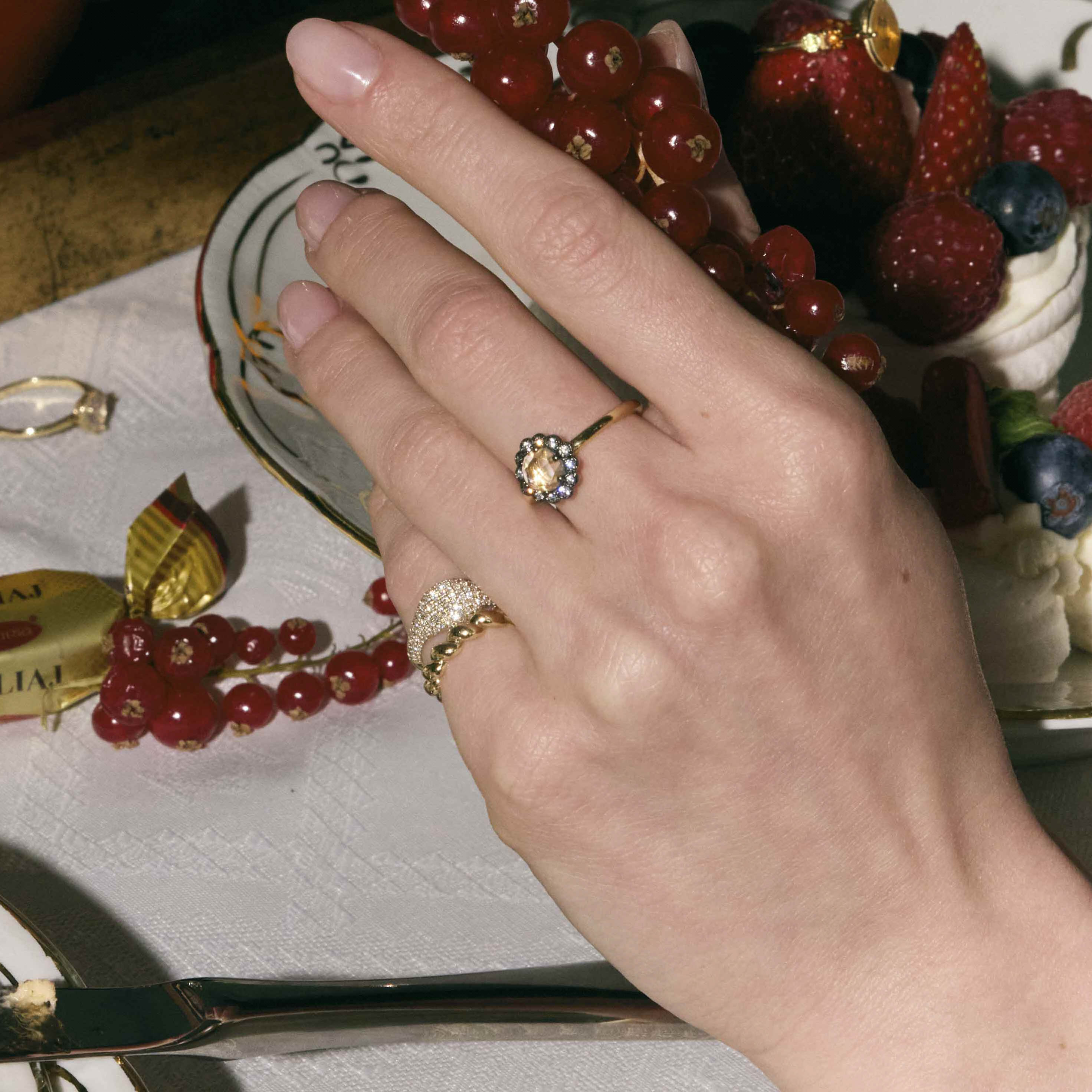 Hand wearing rings with diamonds, and pinky rings on a table with fruit and jewelry.