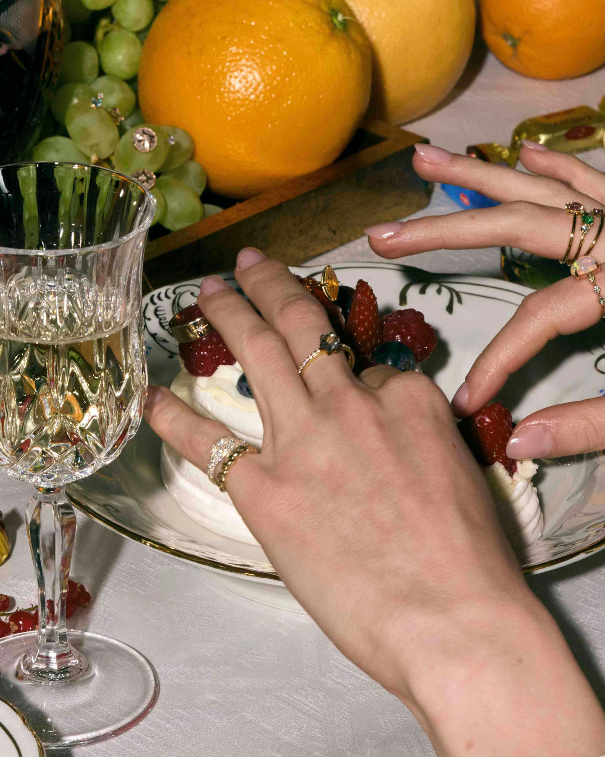Close-up of hands wearing gold rings with diamonds around a dessert plate with fruit and a glass of white wine on a table.