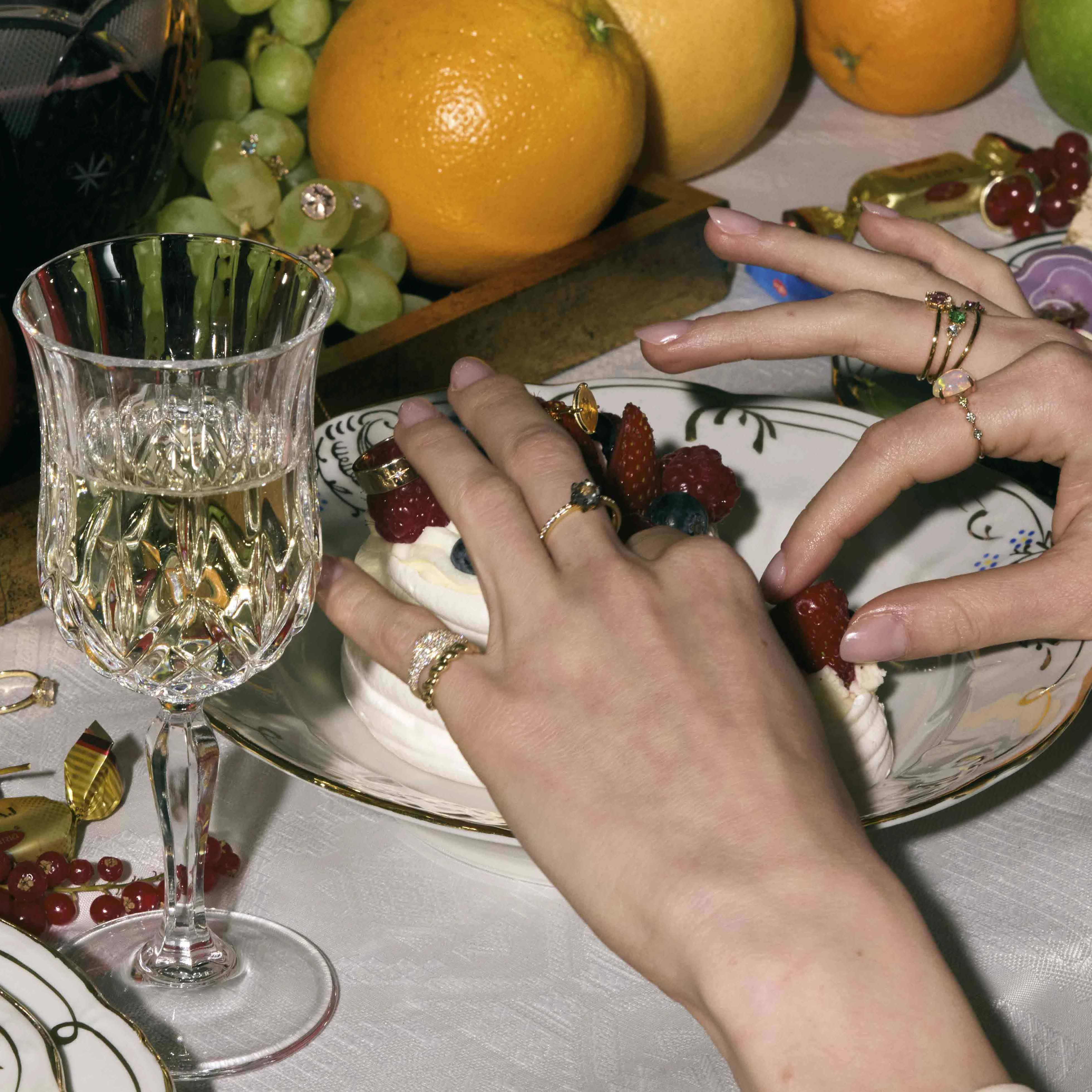 Close-up of hands wearing gold rings with diamonds around a dessert plate with fruit and a glass of white wine on a table.
