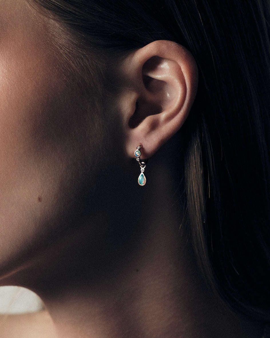 Close-up of an ear with a teardrop earring accessory, with australian opal stone and a diamond accent, against a neutral background.