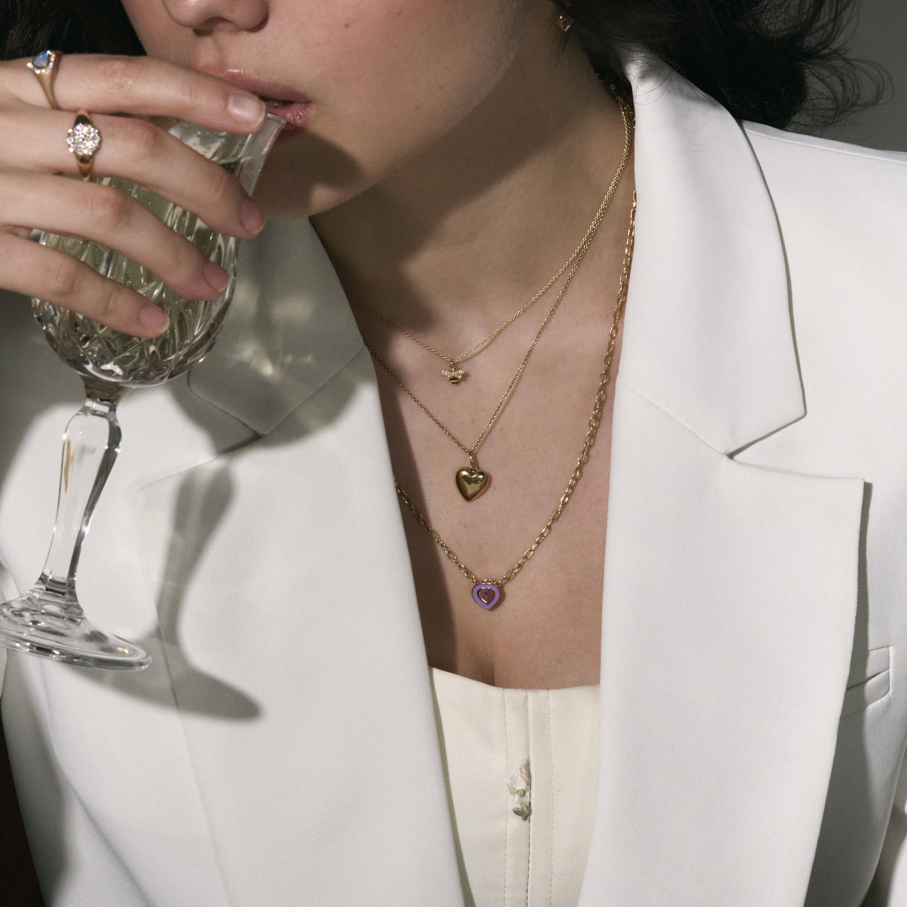 Person wearing gold necklaces with heart and flower pendants, holding a glass.