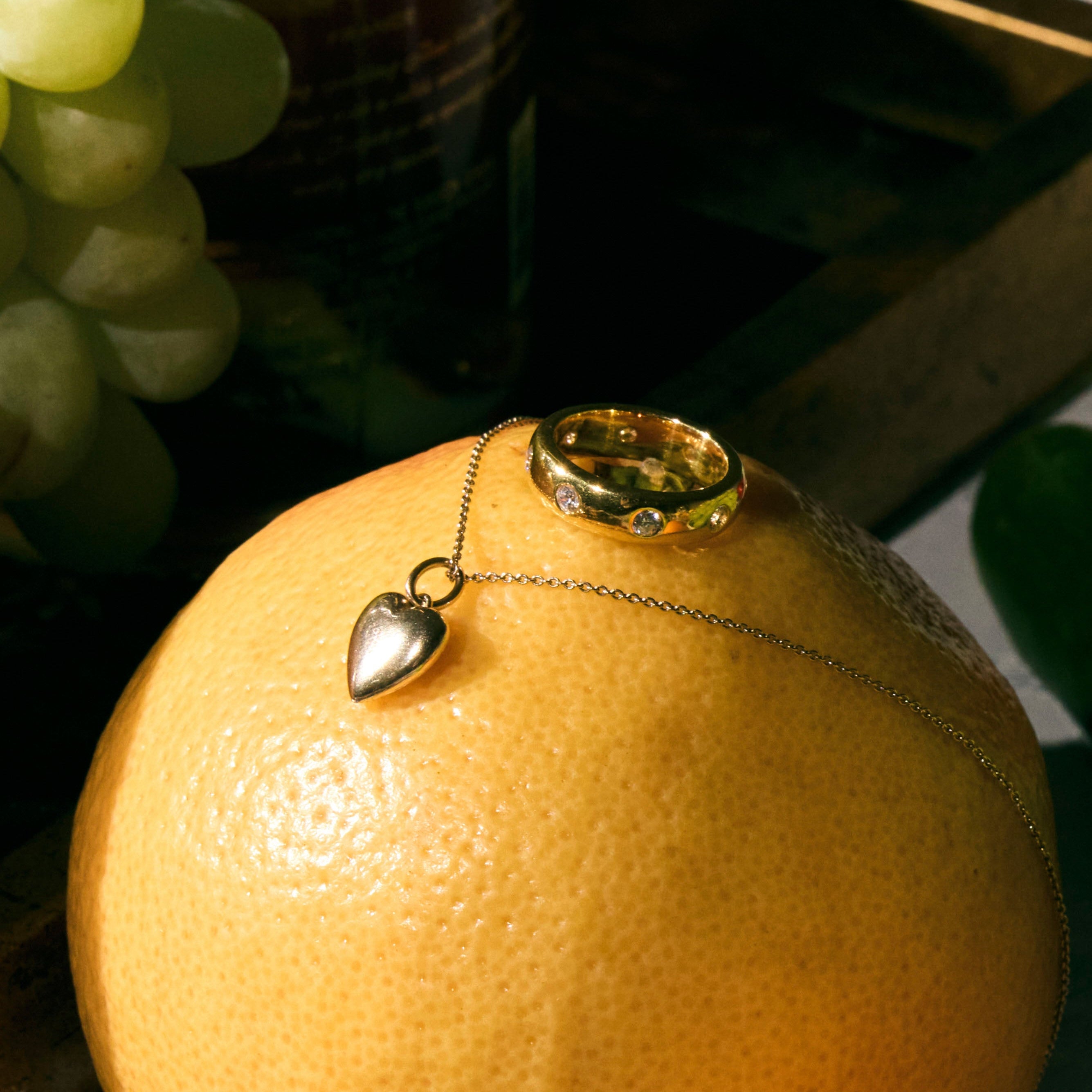 Necklace with a heart pendant, and a cigar band ring with round diamonds on an orange surrounded by green grapes.