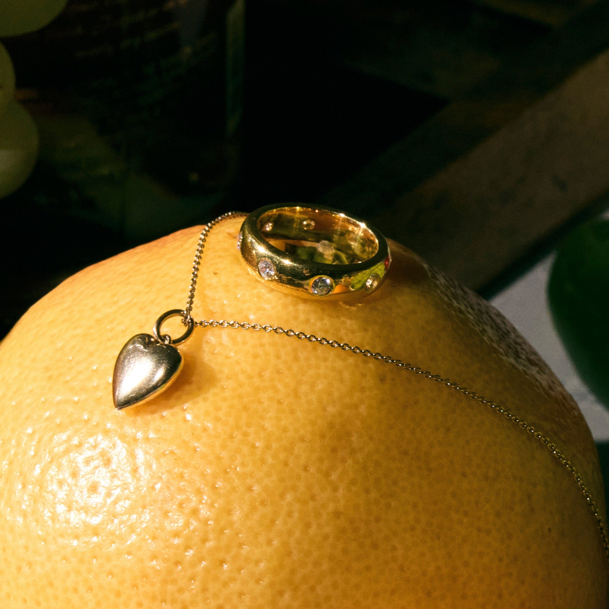 Necklace with a heart pendant, and a cigar band ring with round diamonds on an orange surrounded by green grapes.