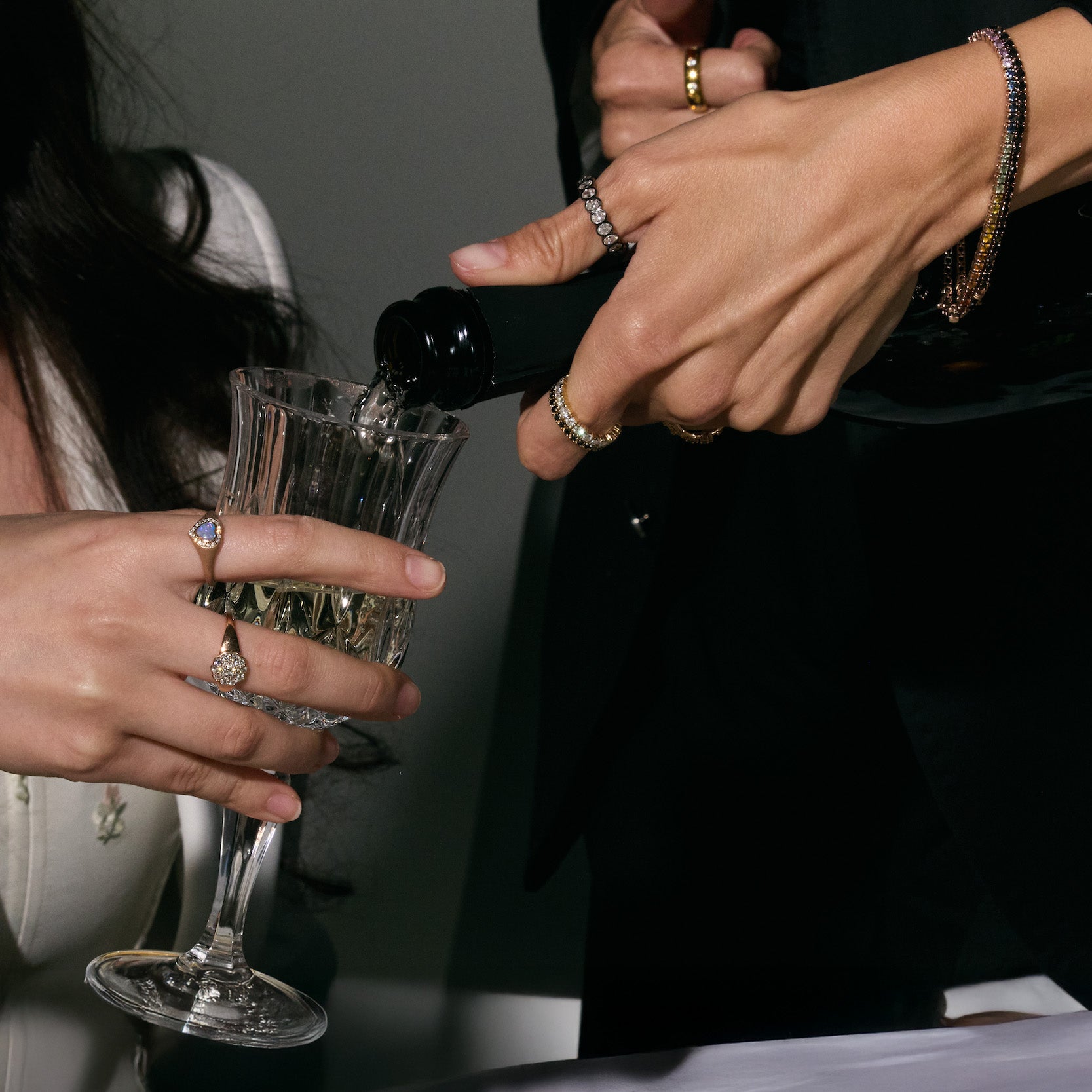 Person pouring champagne into a glass held by another person. Both are wearing gorgeous jewelry, eternity rings and bracelets, cluster diamonds ring and australian opal heart ring