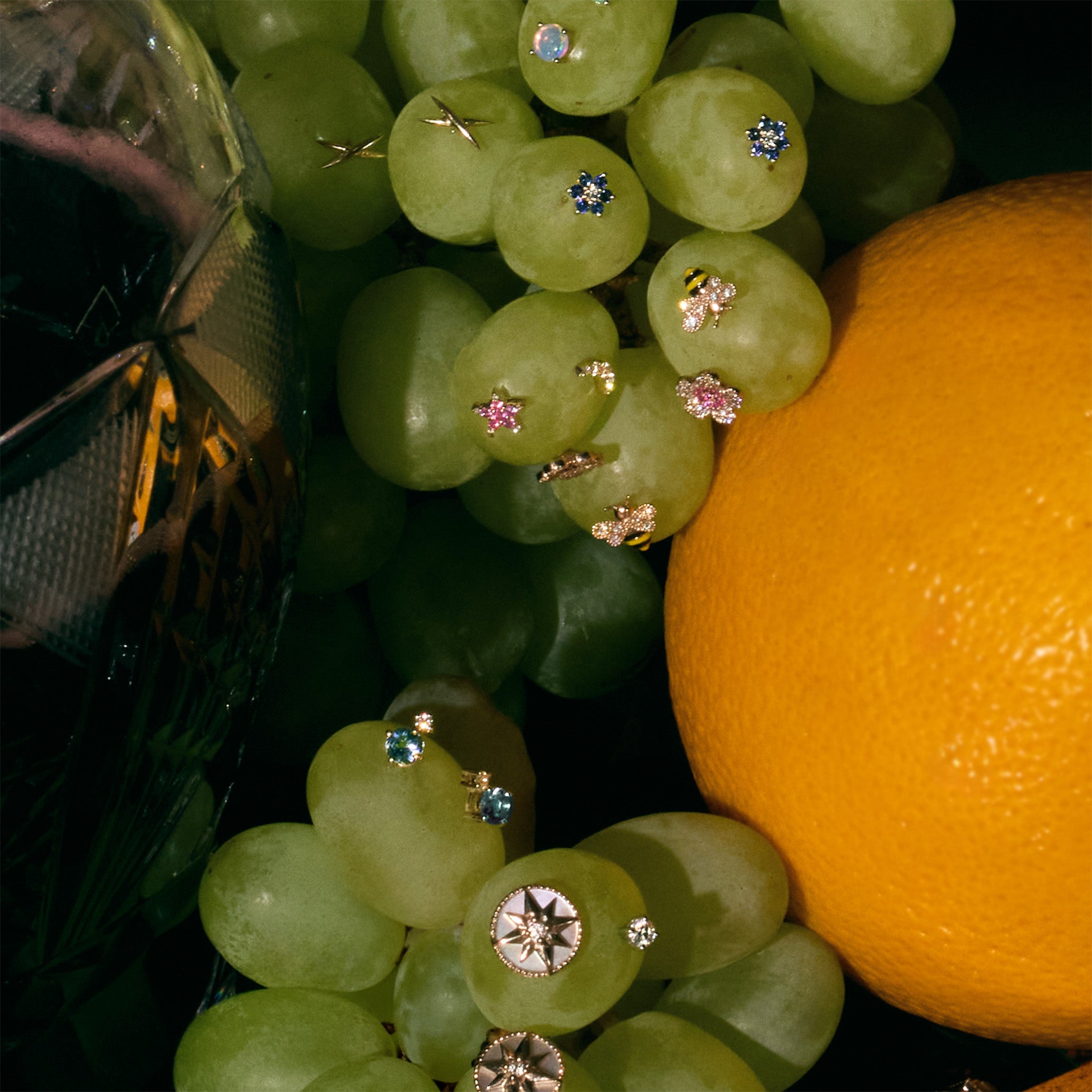 Close-up of green grapes with different stud earrings models piercing the grapes and an orange on a dark background