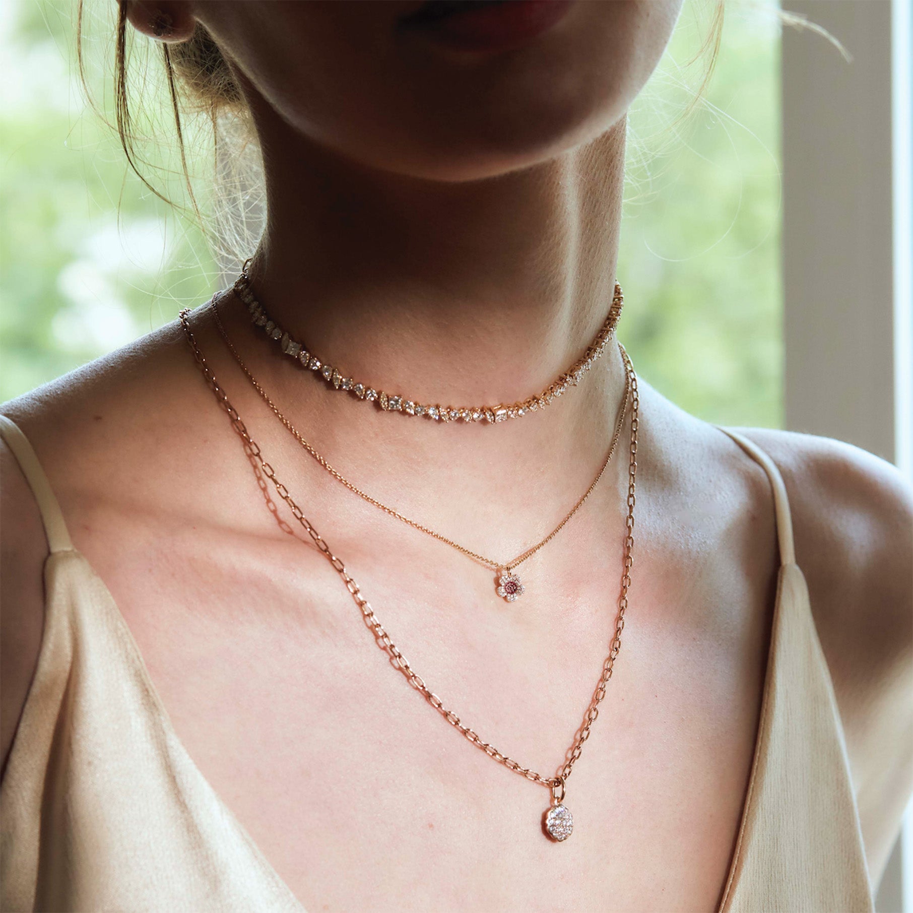 Close-up of a person wearing three rose gold necklaces withe diamonds and a diamond flower pendant, with a blurred natural background