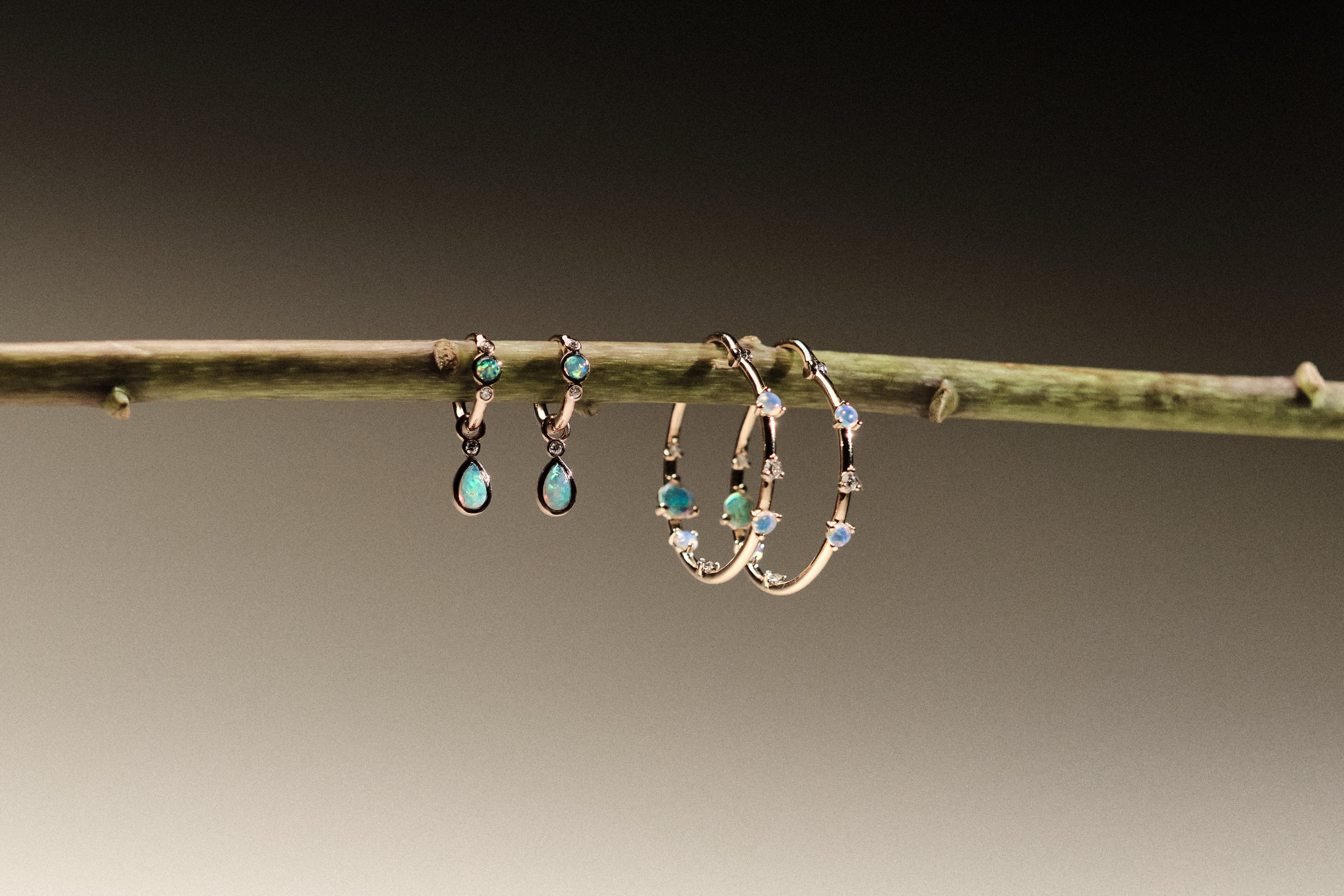 Two pairs of earrings made of 14k gold, lab diamonds and australian opals, hanging on a branch against a neutral background
