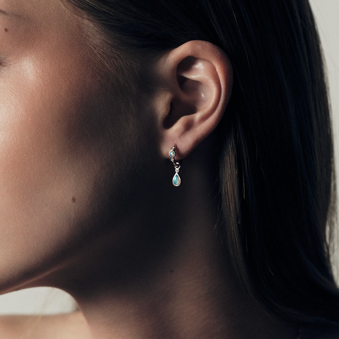 Close-up of an ear with a teardrop earring accessory, with australian opal stone and a diamond accent, against a neutral background.