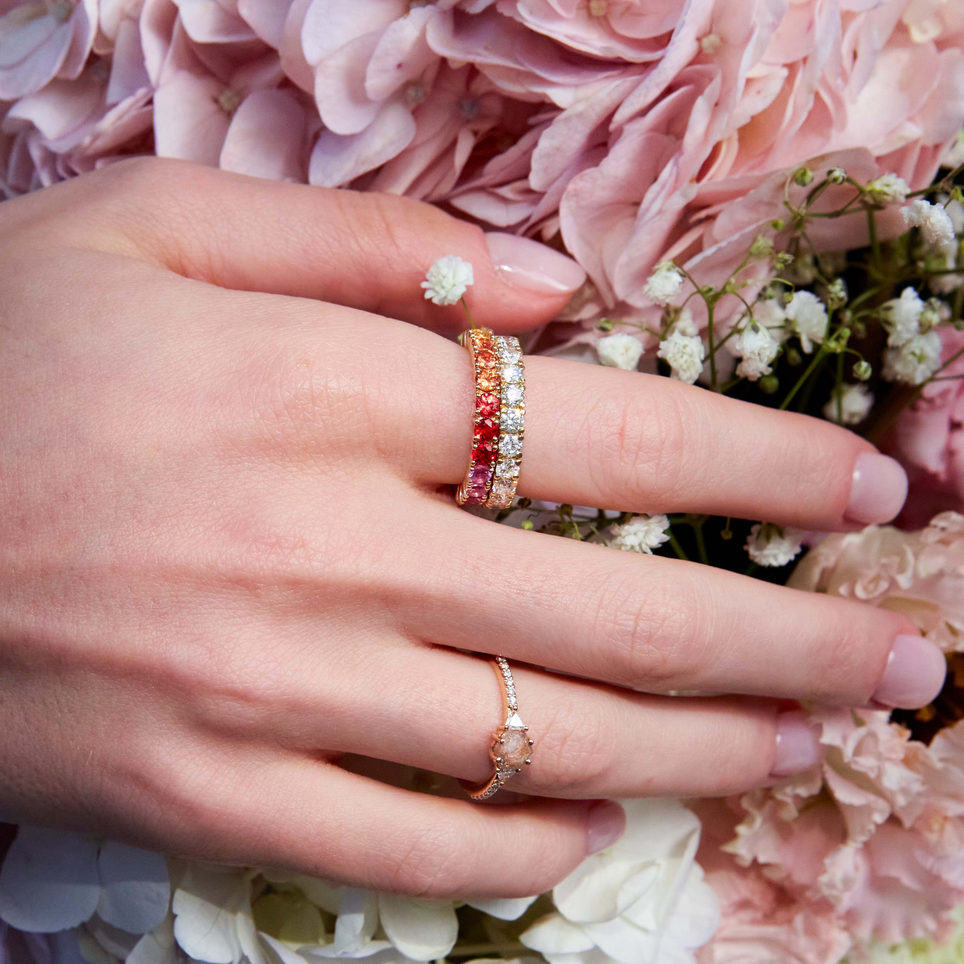 Beautiful eternity rings, one with diamonds and the other with rainbow sapphires, on hand. On the ring finger you can see a dainty salt and pepper diamond engagement ring. The hand is resting on wild colourful flowers. 