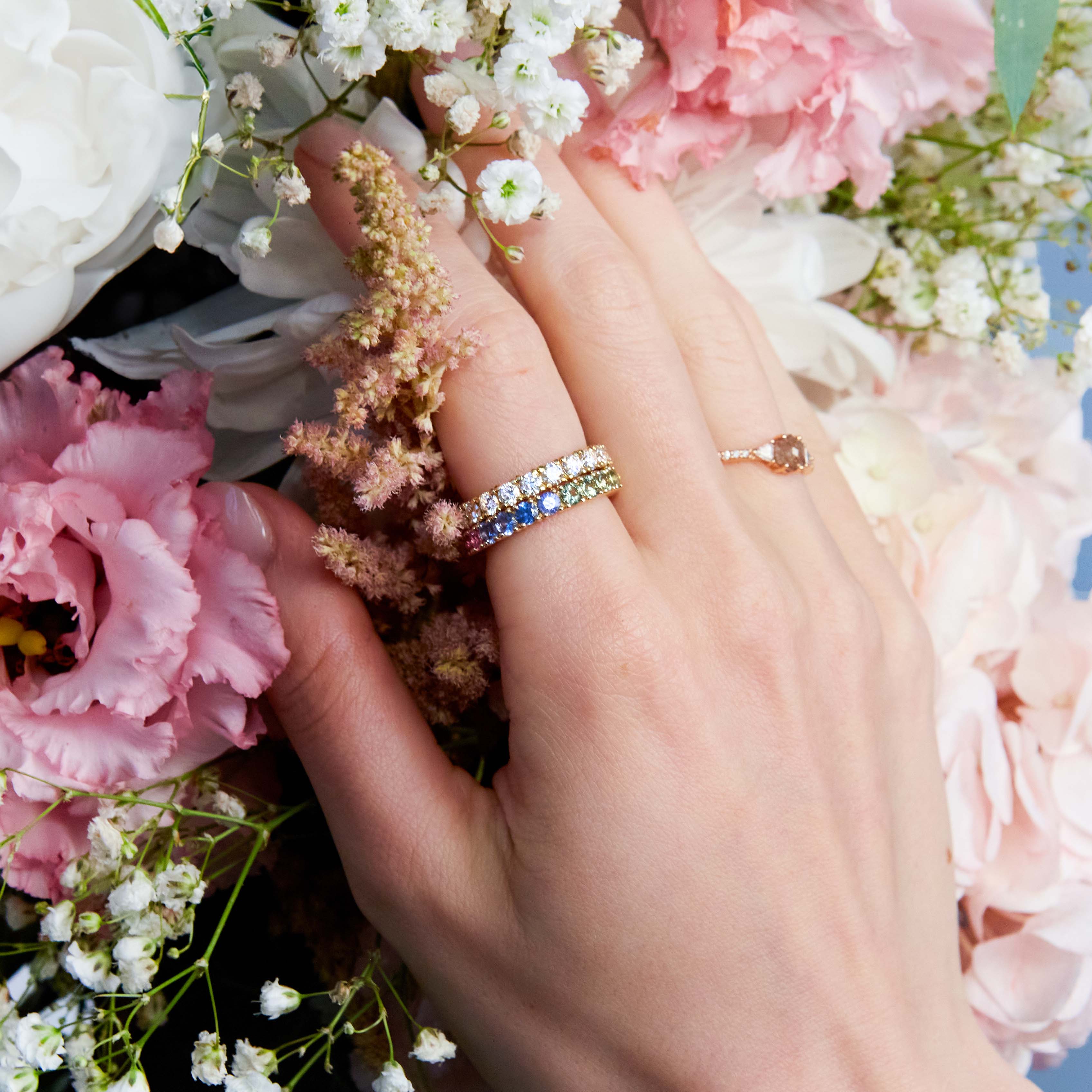 Beautiful eternity rings, one with diamonds and the other with rainbow sapphires on hand. The hand is resting on  wild colourful flowers. 