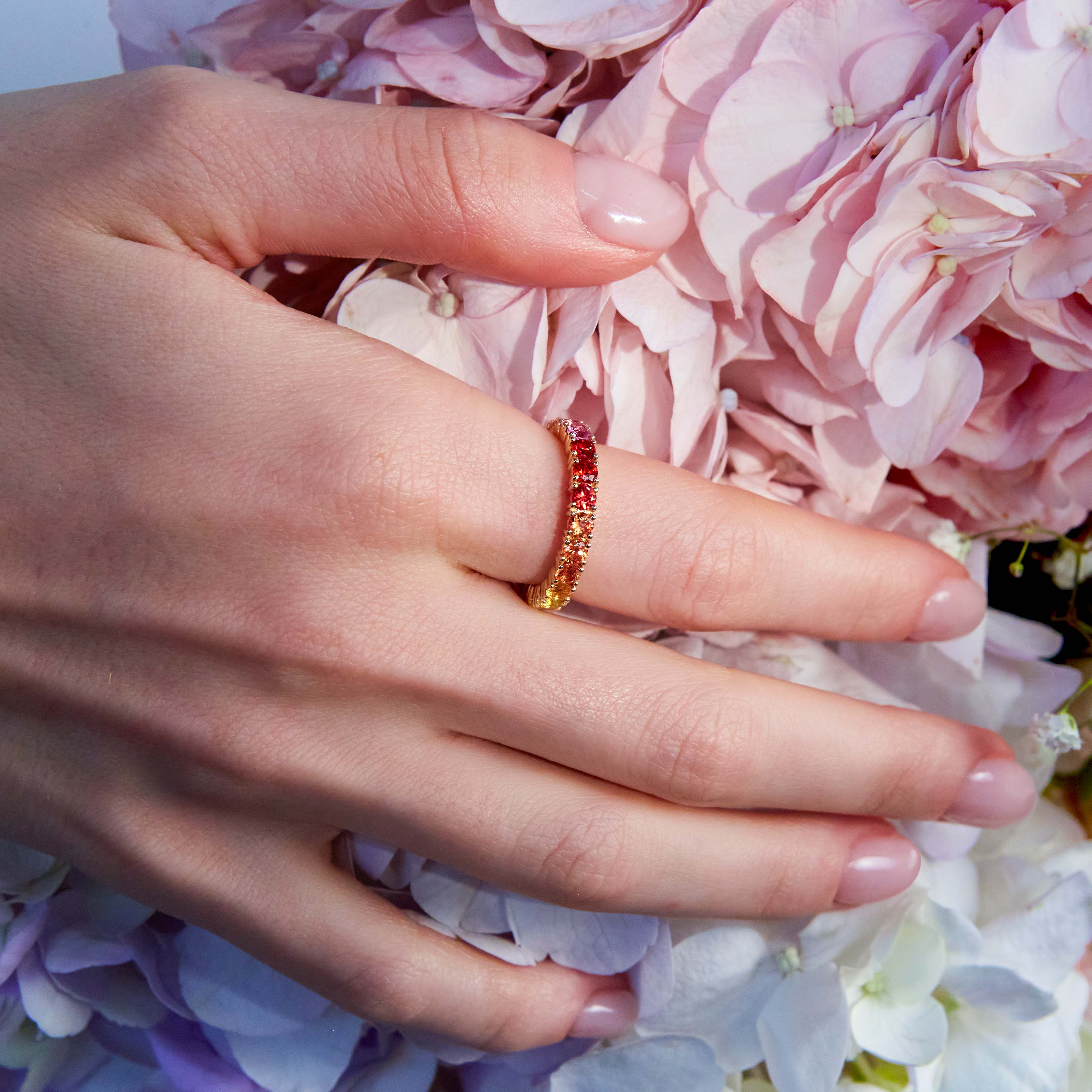 Beautiful eternity rainbow sapphires ring on hand. The hand is resting on wild colourful flowers. 