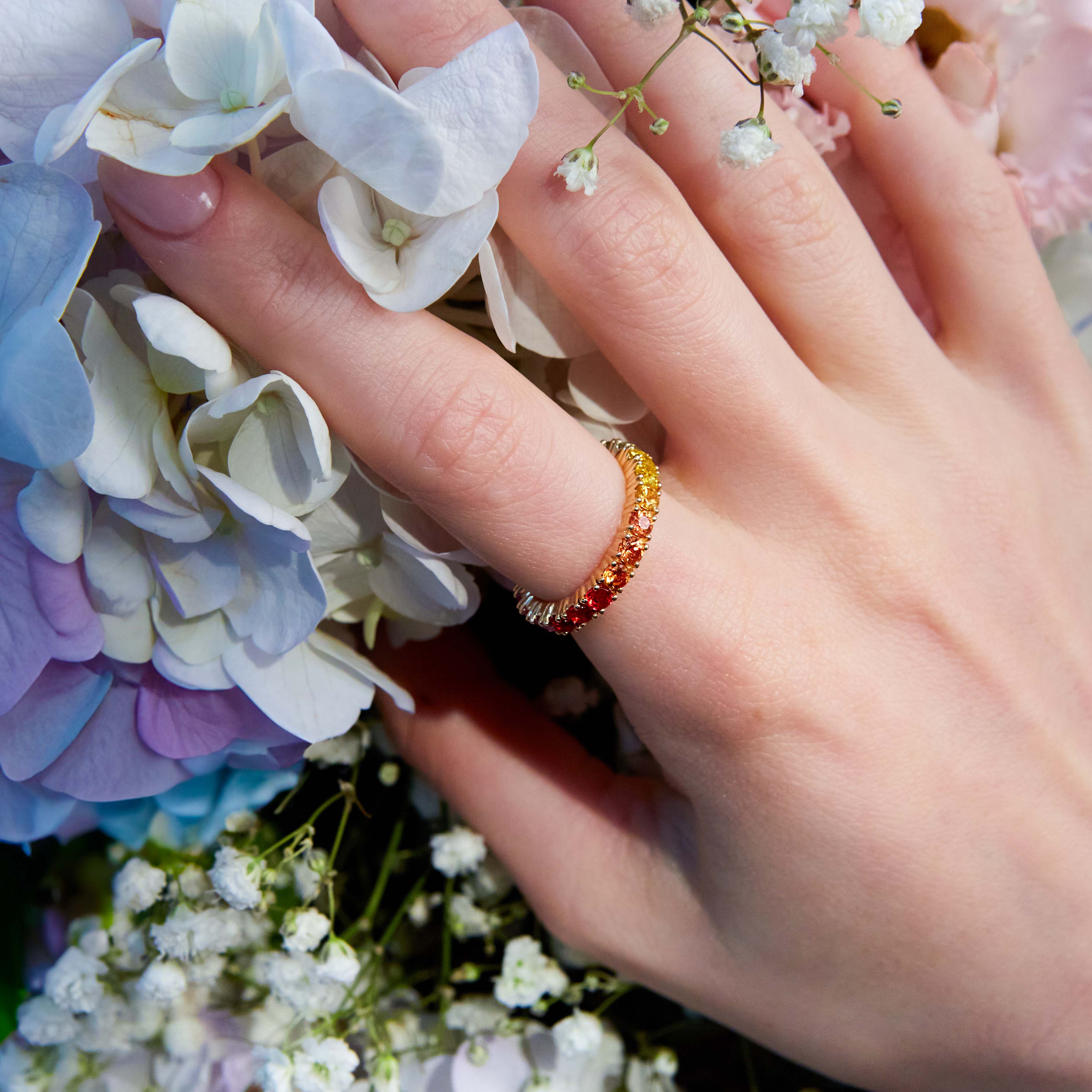 Beautiful eternity rainbow sapphires ring on hand. The hand is resting on a wild colourful flowers. 
