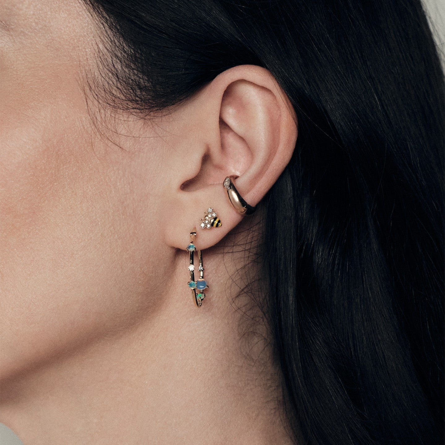 Close-up of a person wearing gold hoop earrings with diamonds and australian opals, baby bee enamela and diamond studs and marquise diamond earcuff.