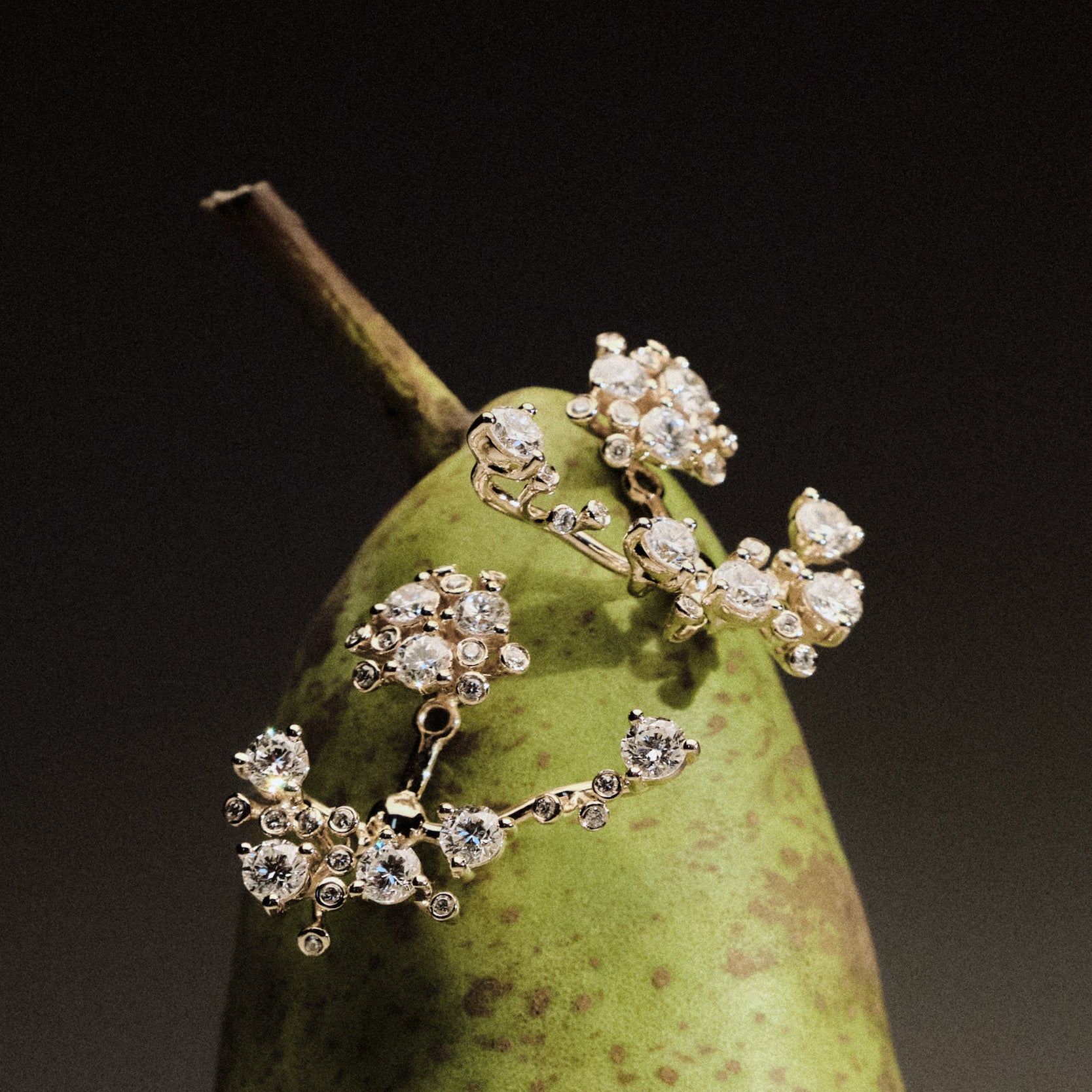 Gold and diamond earrings on a green pear against a dark background.