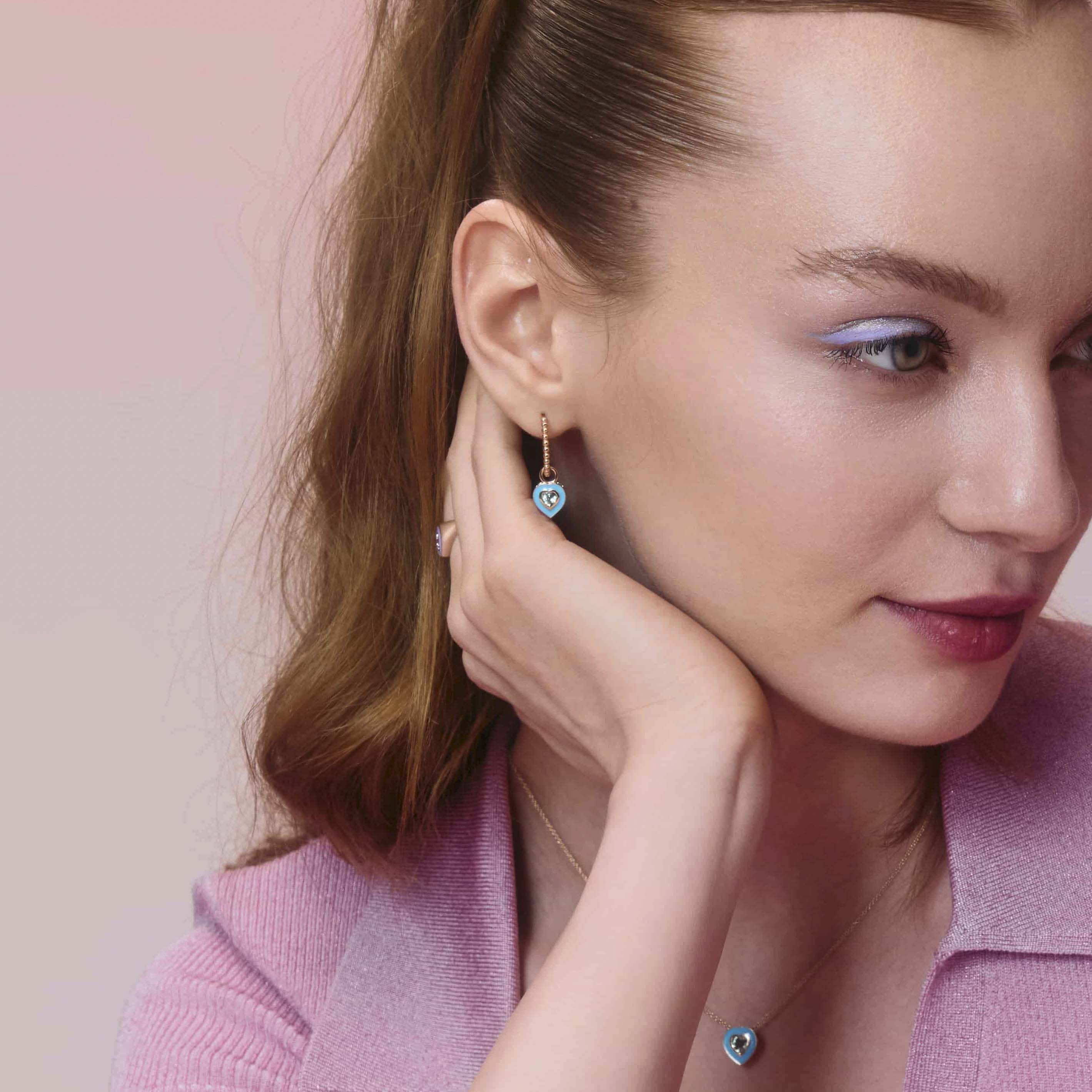  gold hoop earring with a ball design, with enamel heart accessory, on model's ear  displayed against a pink background.