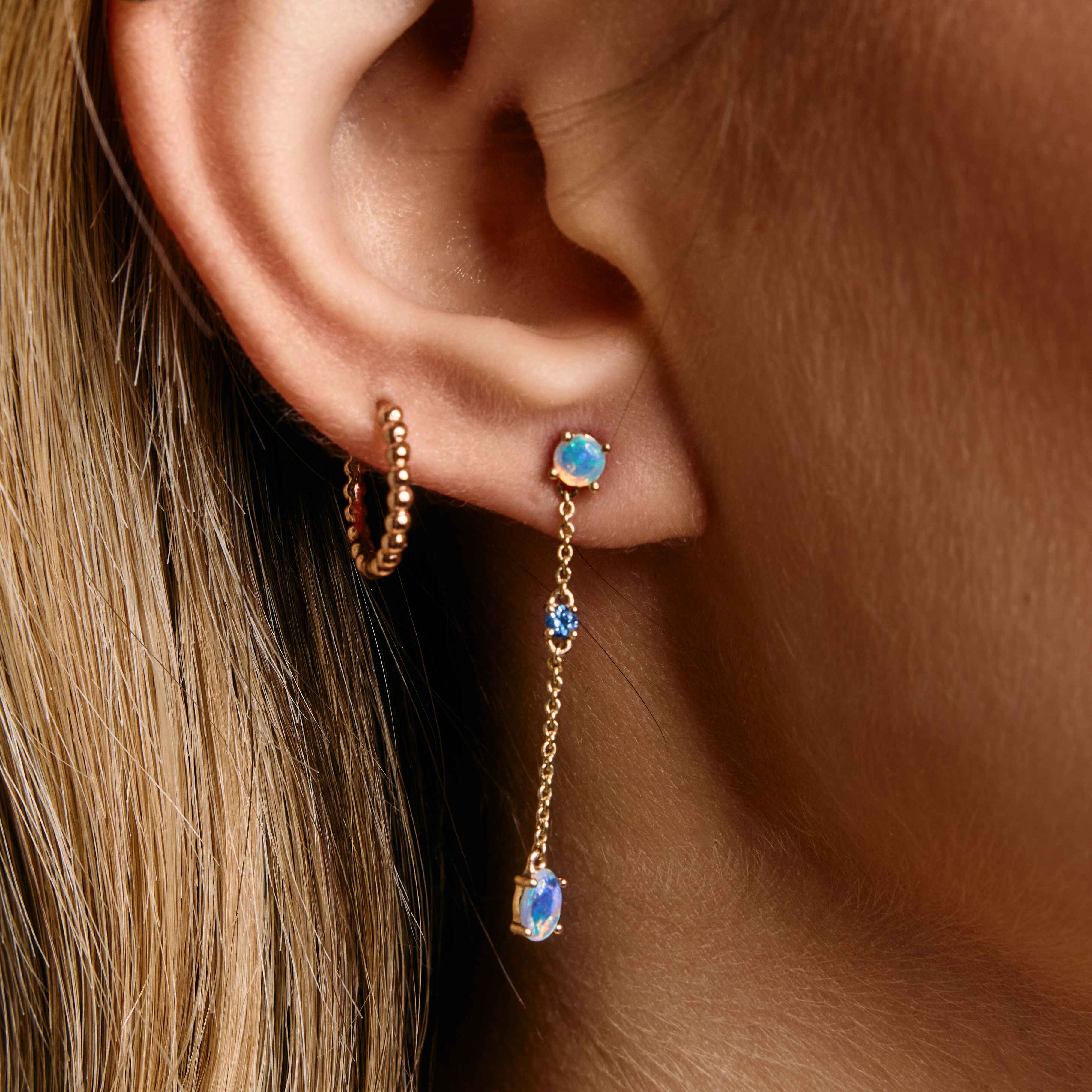 A close-up image of a woman's ear wearing a rose gold, chain link earring with an Australian Opal and a green sapphire.