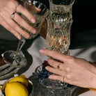 Two hands with gold rings with precious stones holding crystal glasses on a table with fruit and plates.
