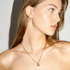 Woman wearing a gold letter necklace with sprinkled diamonds and earring against a neutral background
