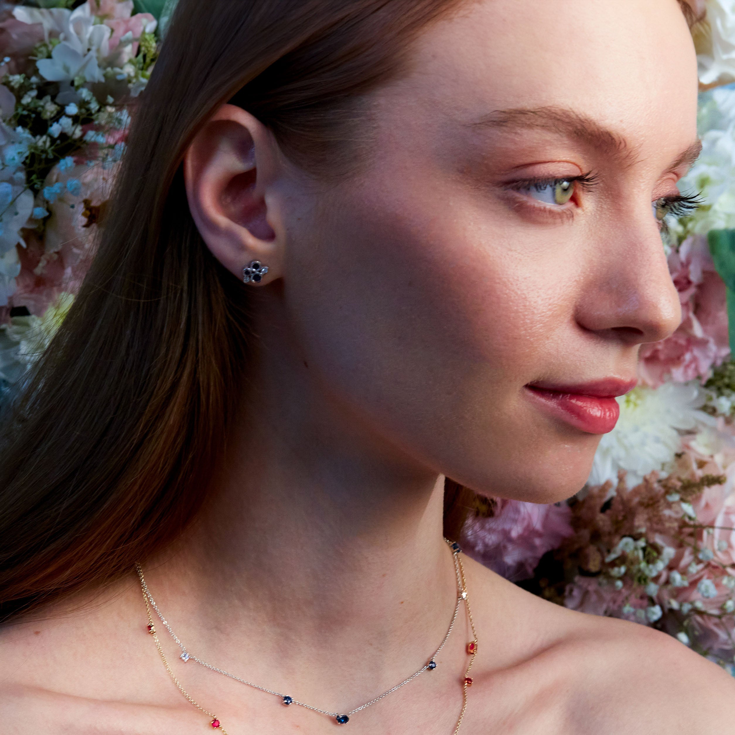 A close-up image of a woman's ear wearing a gold stud earring with a blue sapphire and white diamond cluster design.