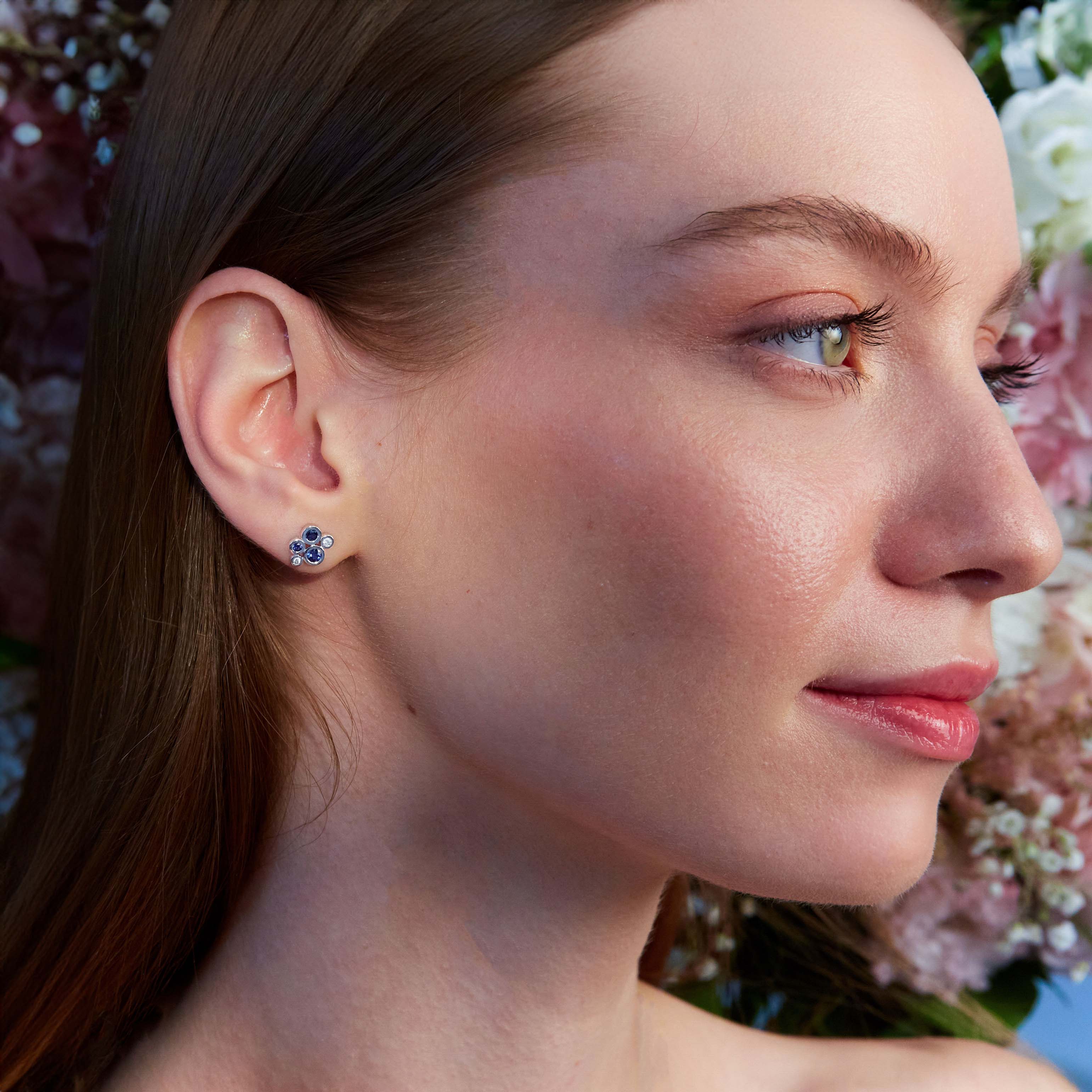 A close-up image of a woman's ear wearing a gold stud earring with a blue sapphire and white diamond cluster design.