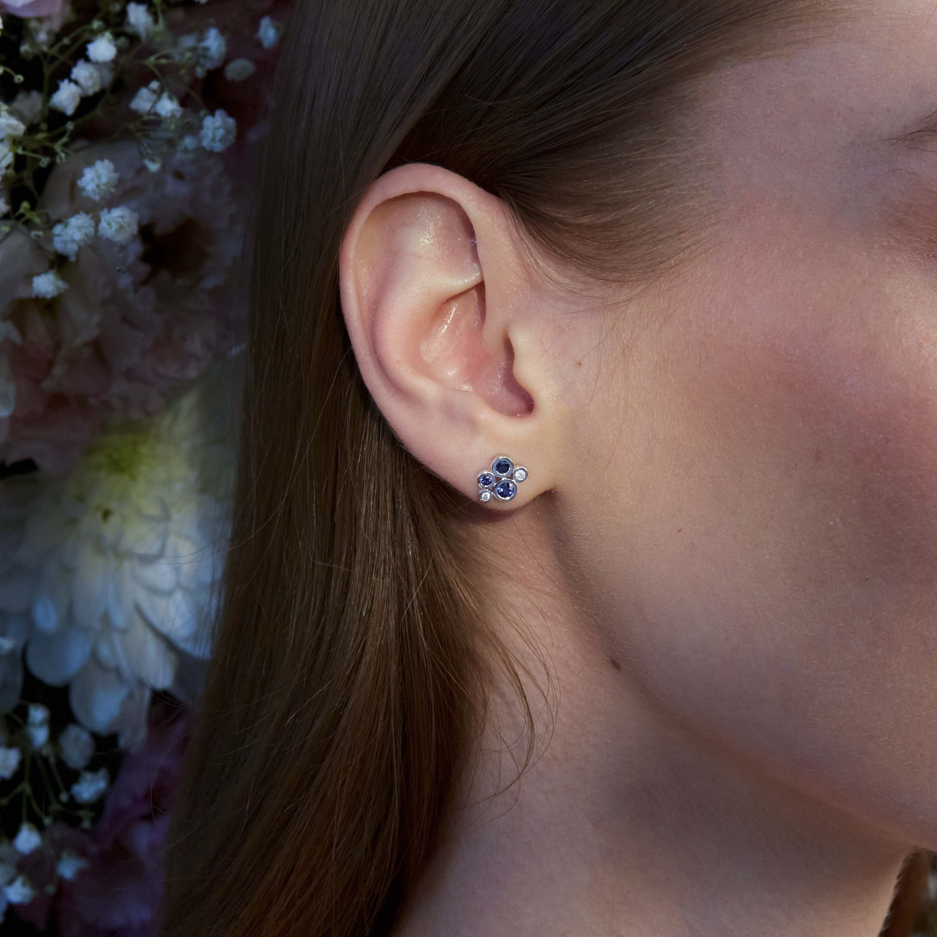 A close-up image of a woman's ear wearing a gold stud earring with a blue sapphire and white diamond cluster design.