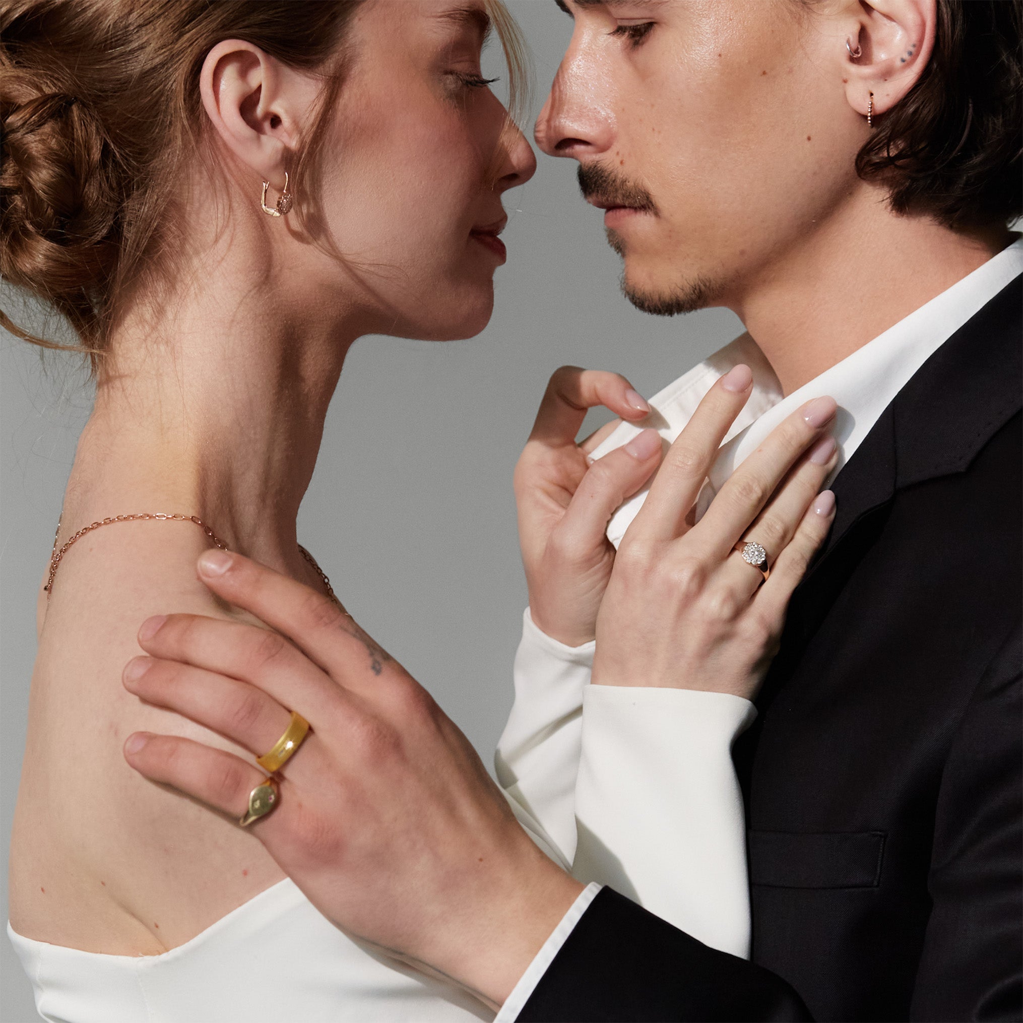 Close-up of a couple embracing with a gray background, They are both wearing diamonds and gold jewelry. 