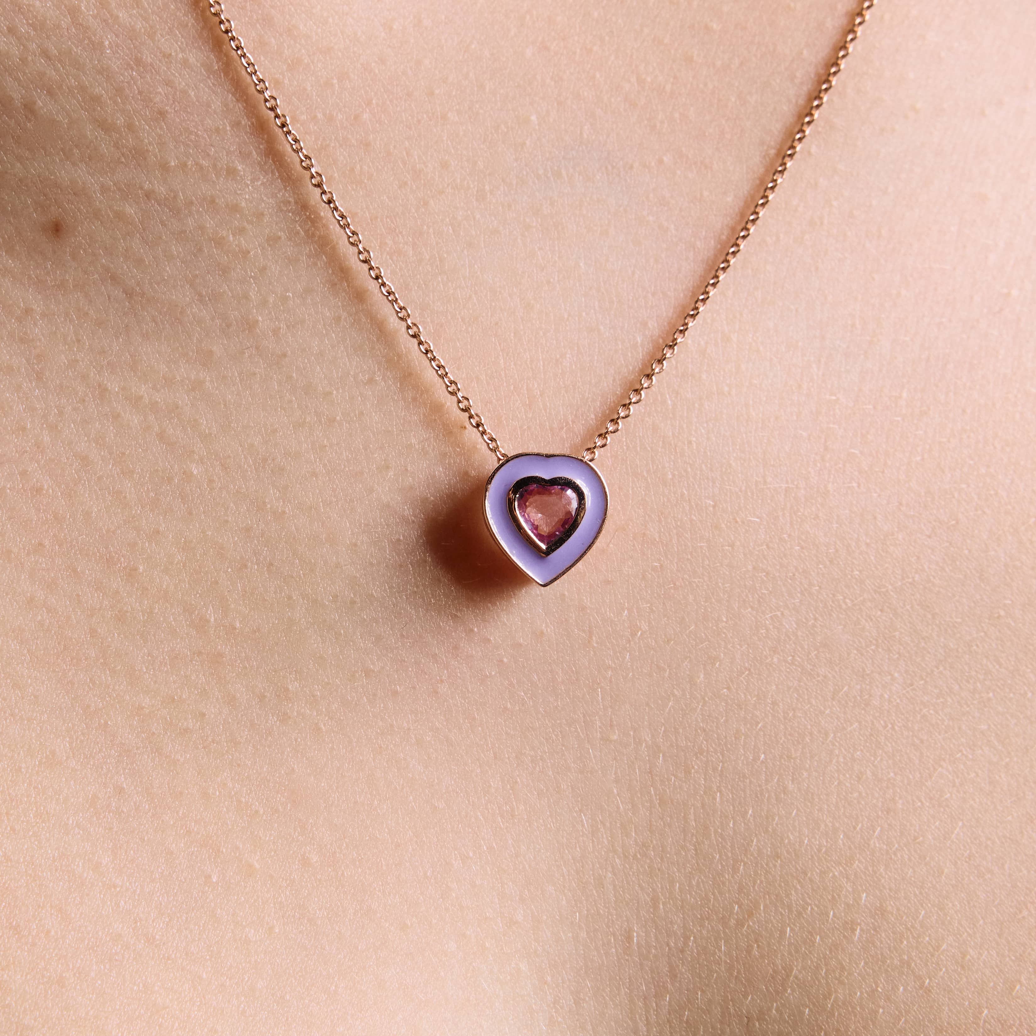 A close-up image of a person wearing a necklaces, with a purple colored enamel pendant and a pink sapphire middle heart, made of rose gold