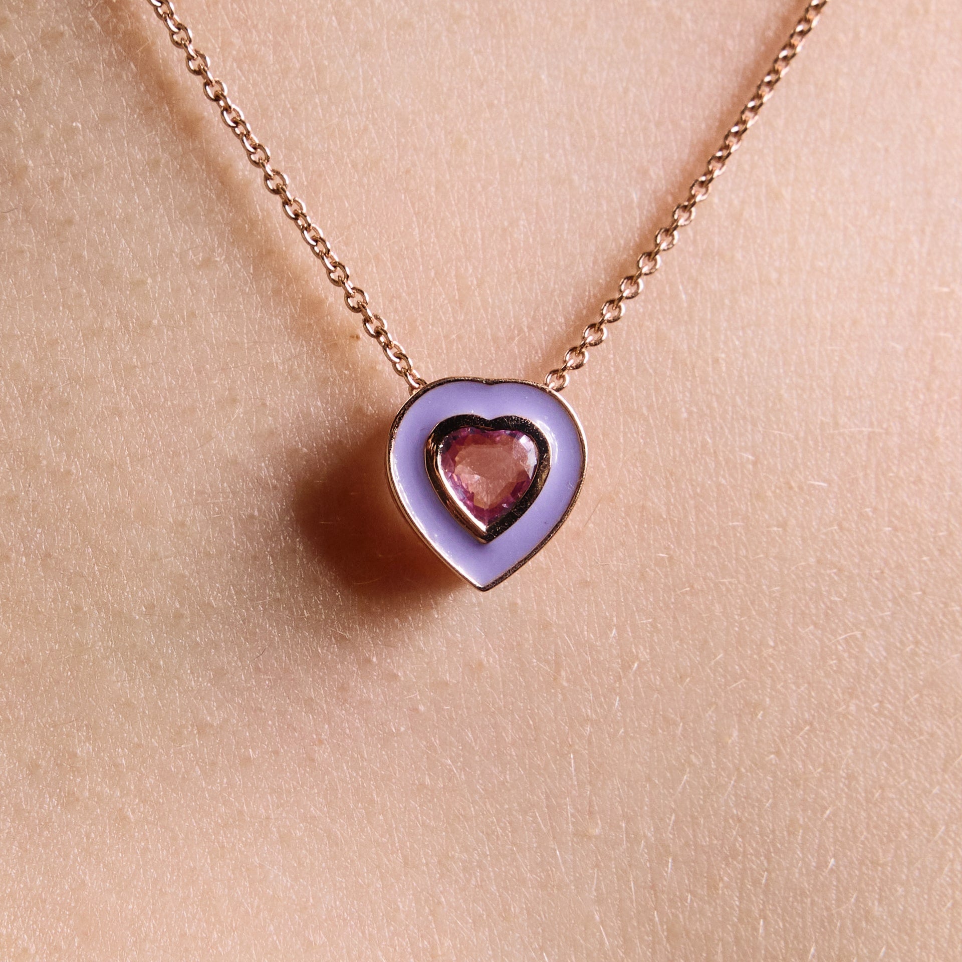 A close-up image of a person wearing a necklaces, with a purple colored enamel pendant and a pink sapphire middle heart, made of rose gold