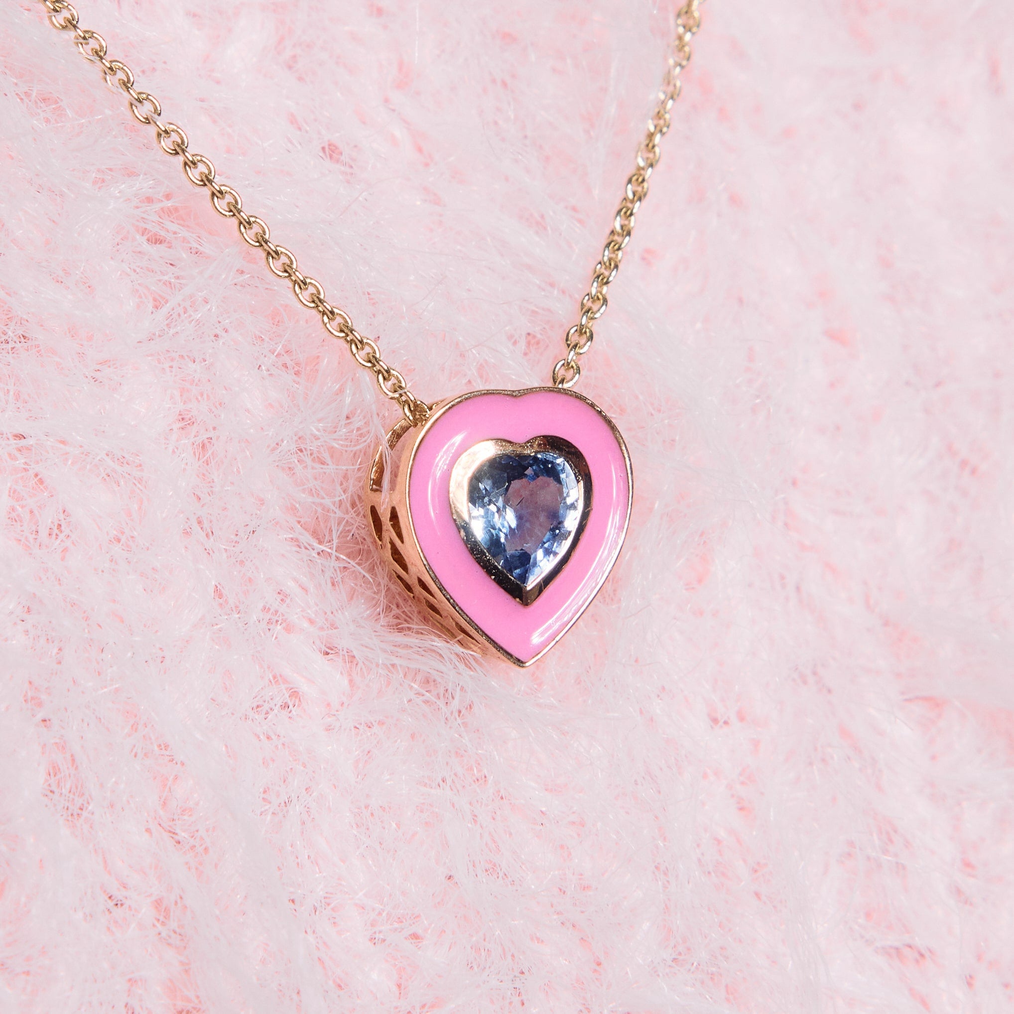 A close-up image of a person wearing a pink coloured enamel pendant, heart shaped, with a blue sapphire heart in the middle