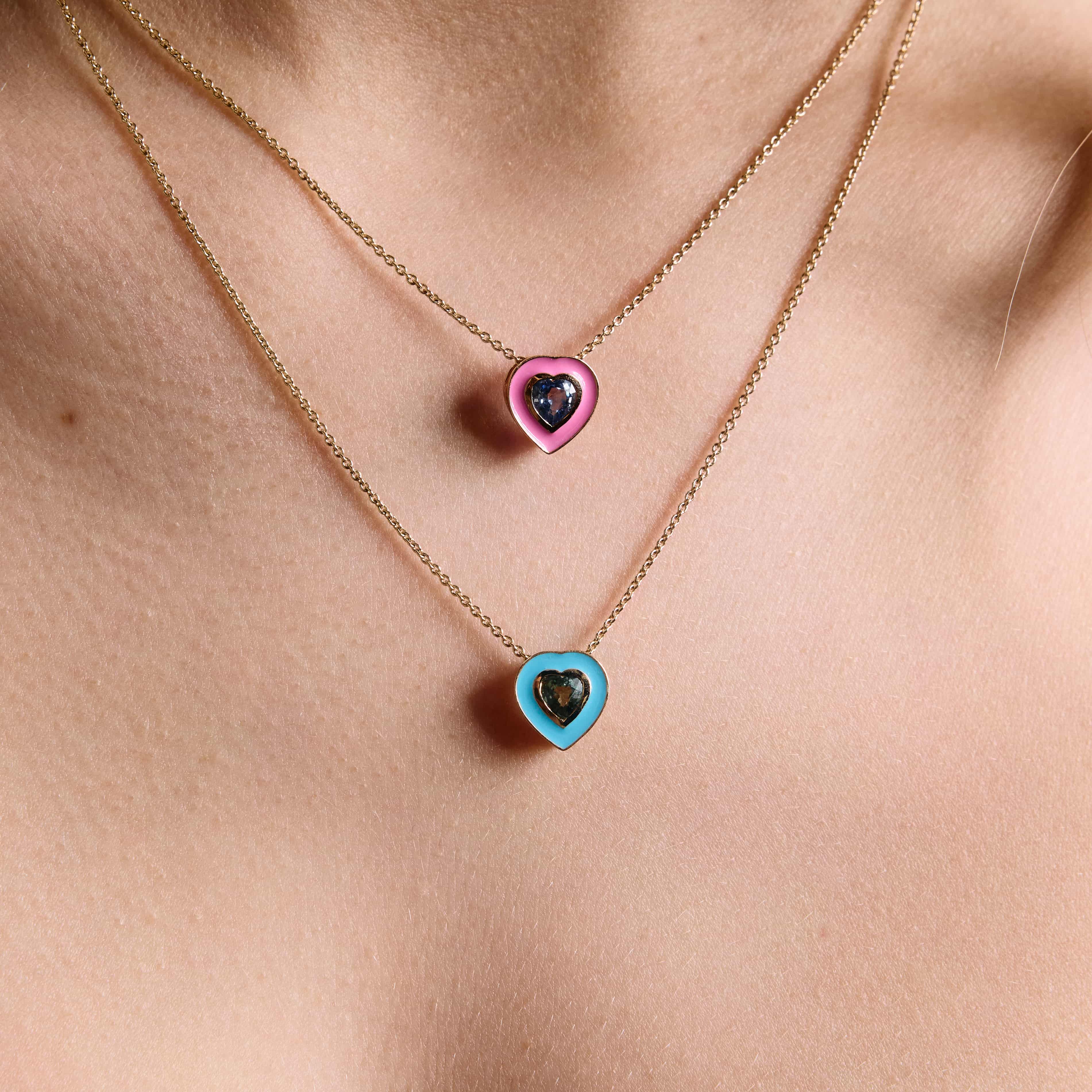 A close-up image of a person wearing two layered necklaces, each with a different colored enamel pendant, showcasing a total of three enamel shades on the pendants.