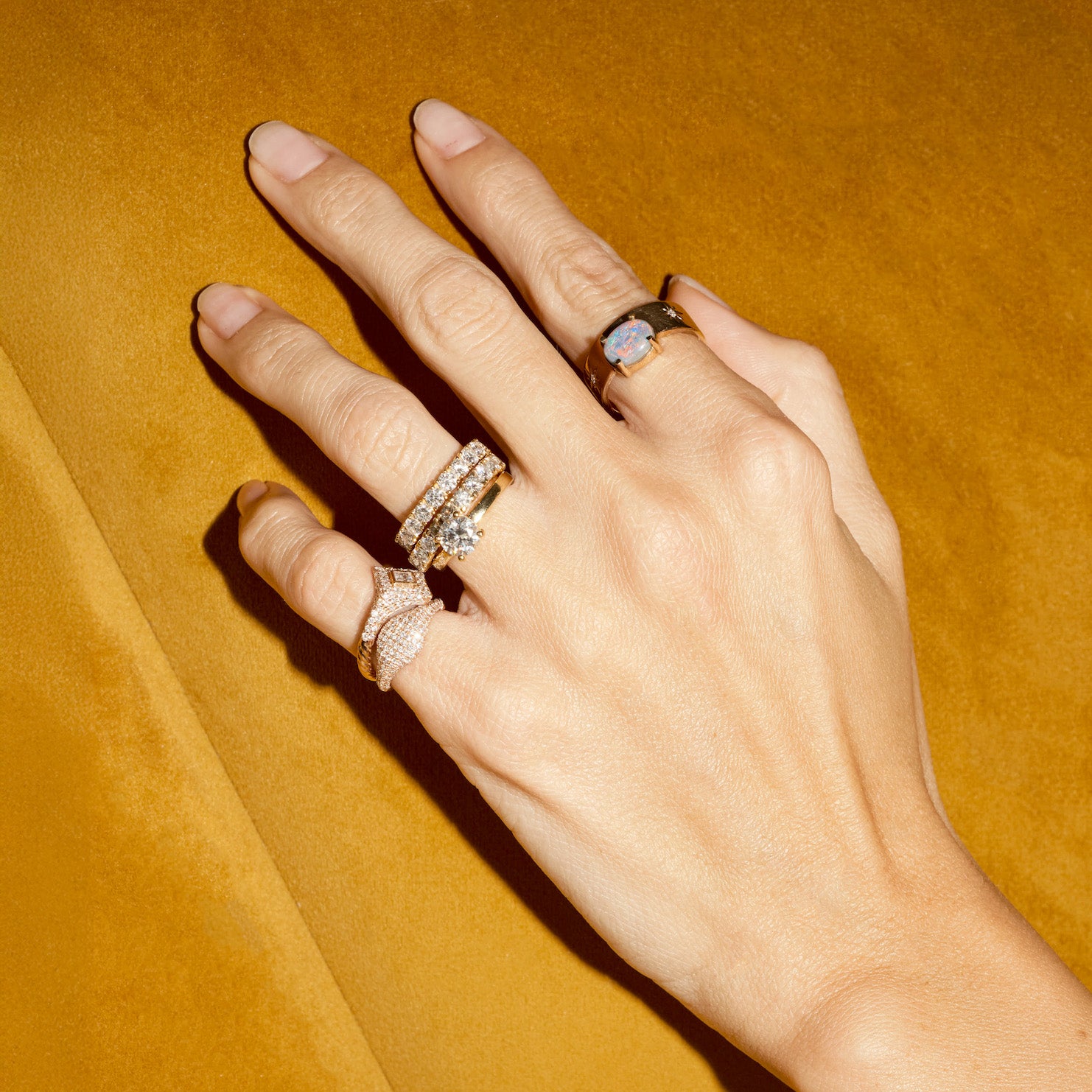 Hand wearing multiple diamond rings, and an australian opal band, on a mustard yellow background