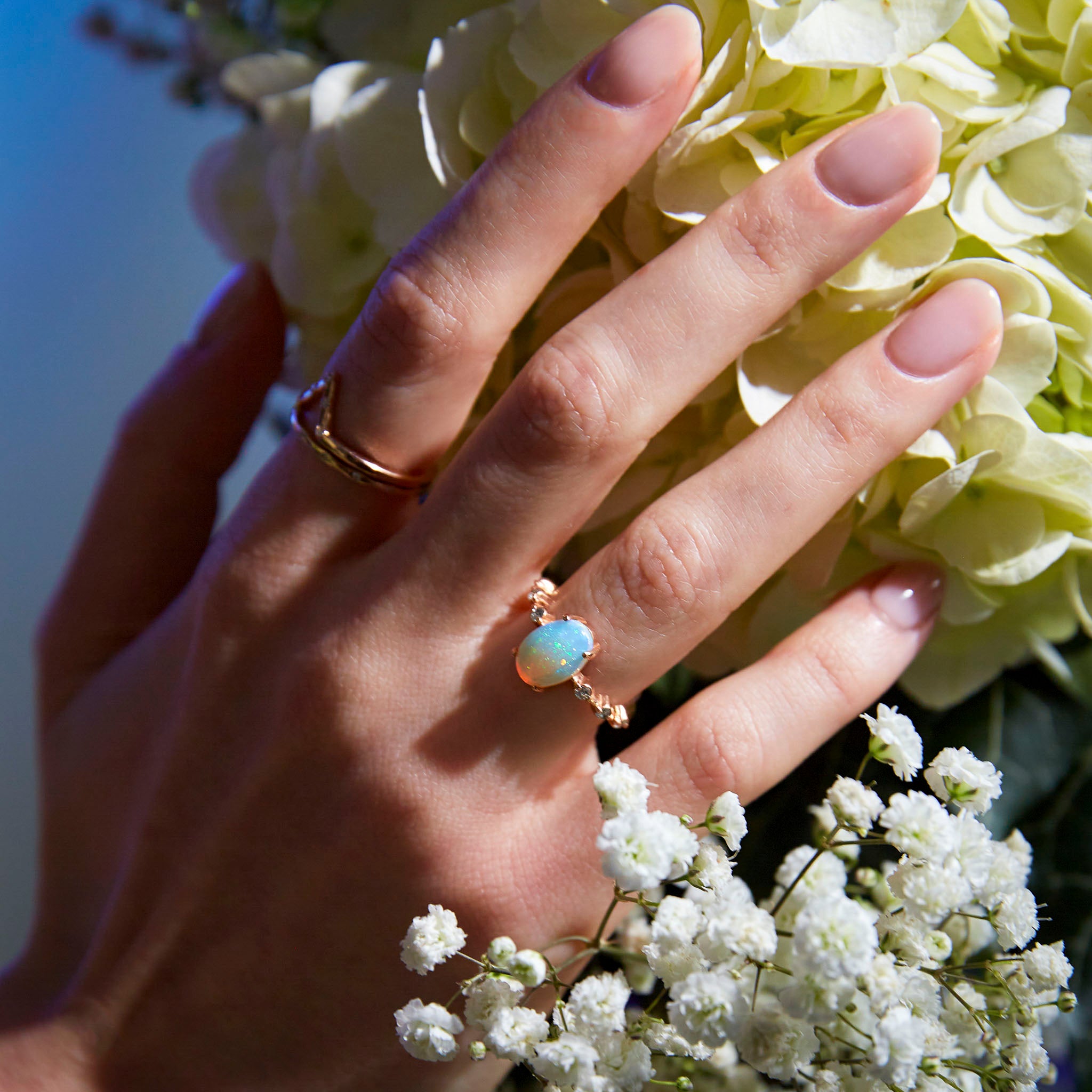 A hand showcasing a gold ring with an opal center stone and white diamond accents on the band, placed on a floral background.