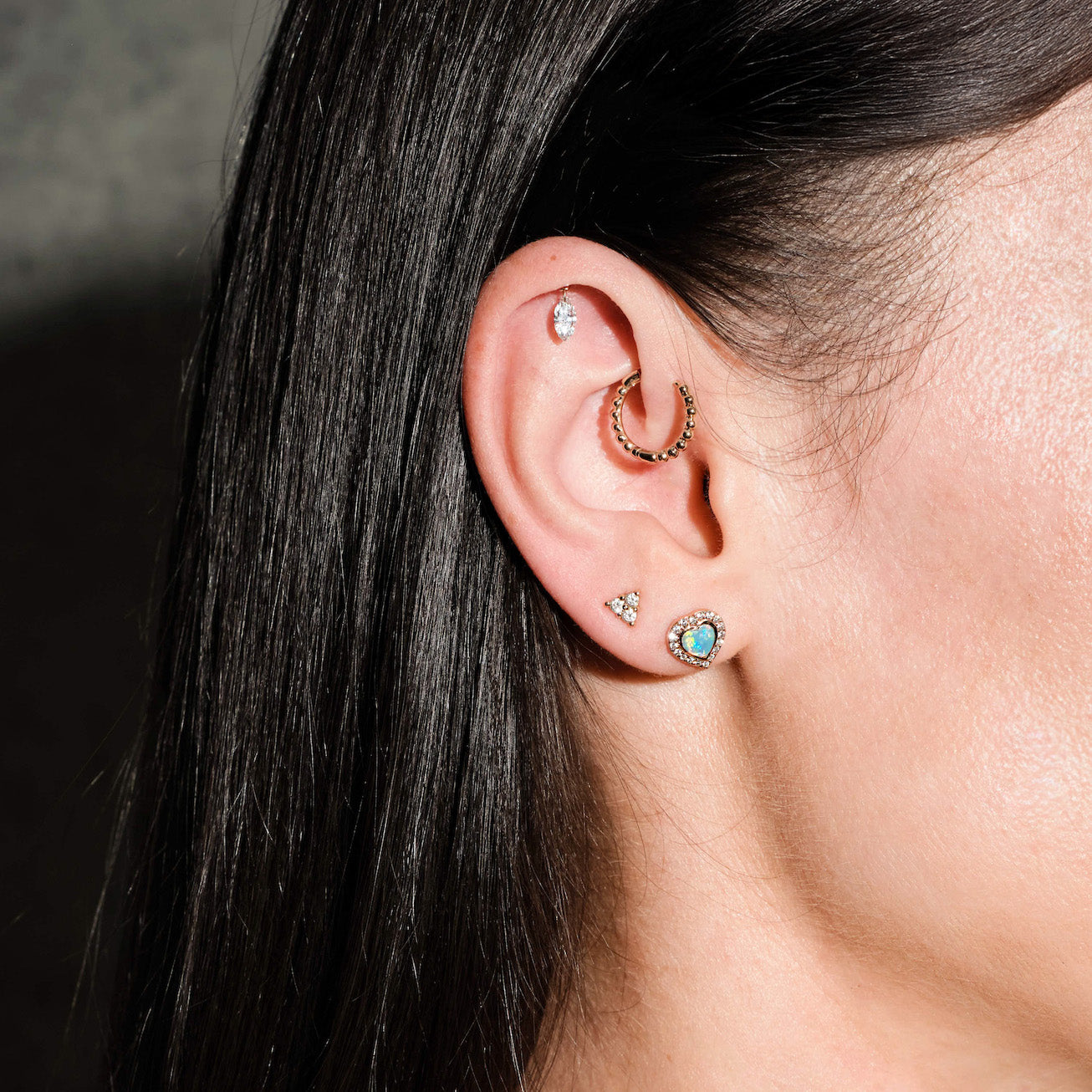 Close-up of an ear with multiple earrings, floating marquise diamond, beaded hoop, diamonds triangle stud, australian opal and diamonds heart studs, on a dark background
