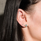Close-up of an ear with multiple earrings, floating marquise diamond, beaded hoop, diamonds triangle stud, australian opal and diamonds heart studs, on a dark background