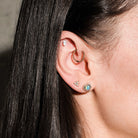 Close-up of an ear with multiple earrings, floating marquise diamond, beaded hoop, diamonds triangle stud, australian opal and diamonds heart studs, on a dark background
