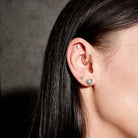 Close-up of an ear with multiple earrings, floating marquise diamond, beaded hoop, diamonds triangle stud, australian opal and diamonds heart studs, on a dark background
