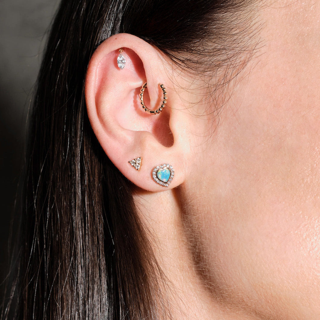 Close-up of an ear with multiple earrings, floating marquise diamond, beaded hoop, diamonds triangle stud, australian opal and diamonds heart studs, on a dark background
