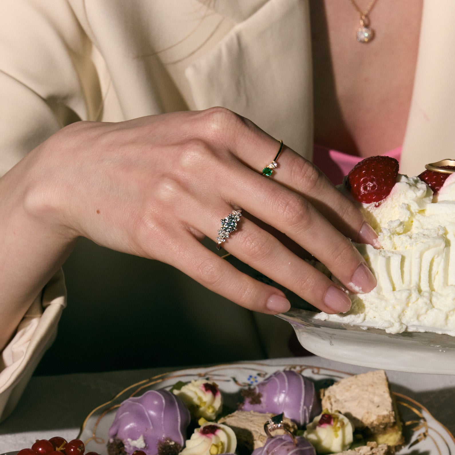 Person wearing two rings, with diamonds and one with emerald and diamond , reaching for a dessert on a plate.